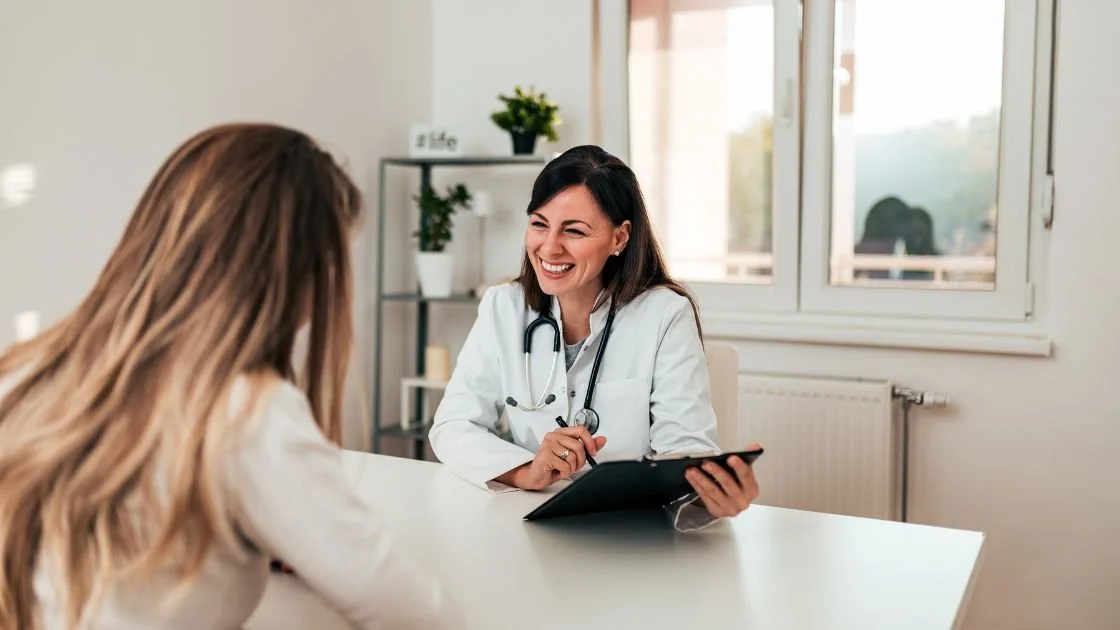 Two female doctors or medical professionals having a conversation in an office, smiling, with medical charts and a window in the background.