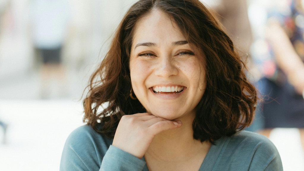 A young woman with shoulder-length curly hair smiling at the camera, wearing a teal long-sleeve shirt, in a bright indoor setting.