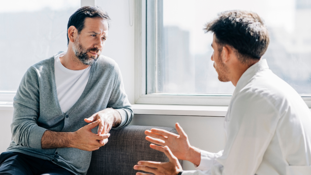 A man and a male doctor having a serious conversation in a bright office, seated on a gray sofa by a window.