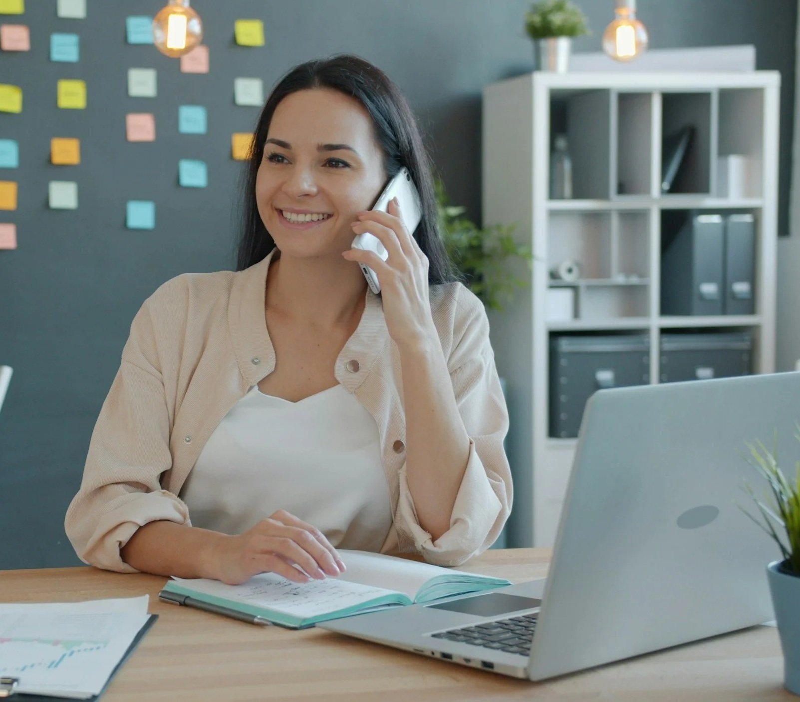 A woman smiling while talking on a landline phone in an office with a laptop and notebooks on the desk.