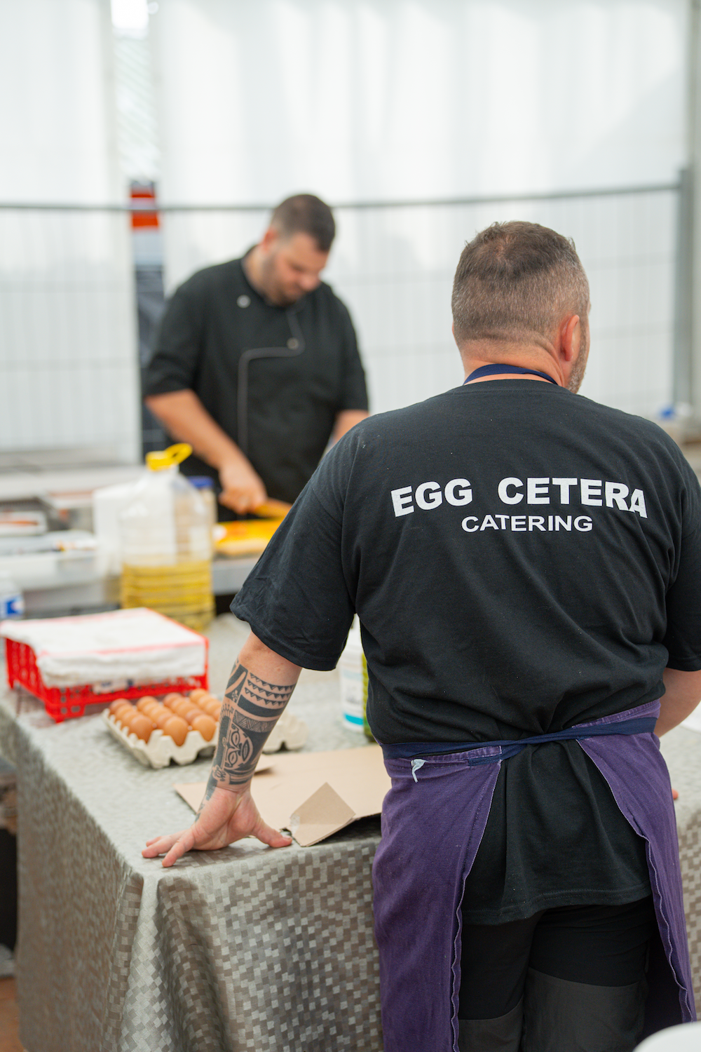 Deux hommes en cuisine, l'un portant un t-shirt noir avec l'inscription "Egg Cetera Catering" vu de dos, et l'autre en arrière-plan, préparant des ingrédients sur une table, avec des œufs et d'autres fournitures culinaires.