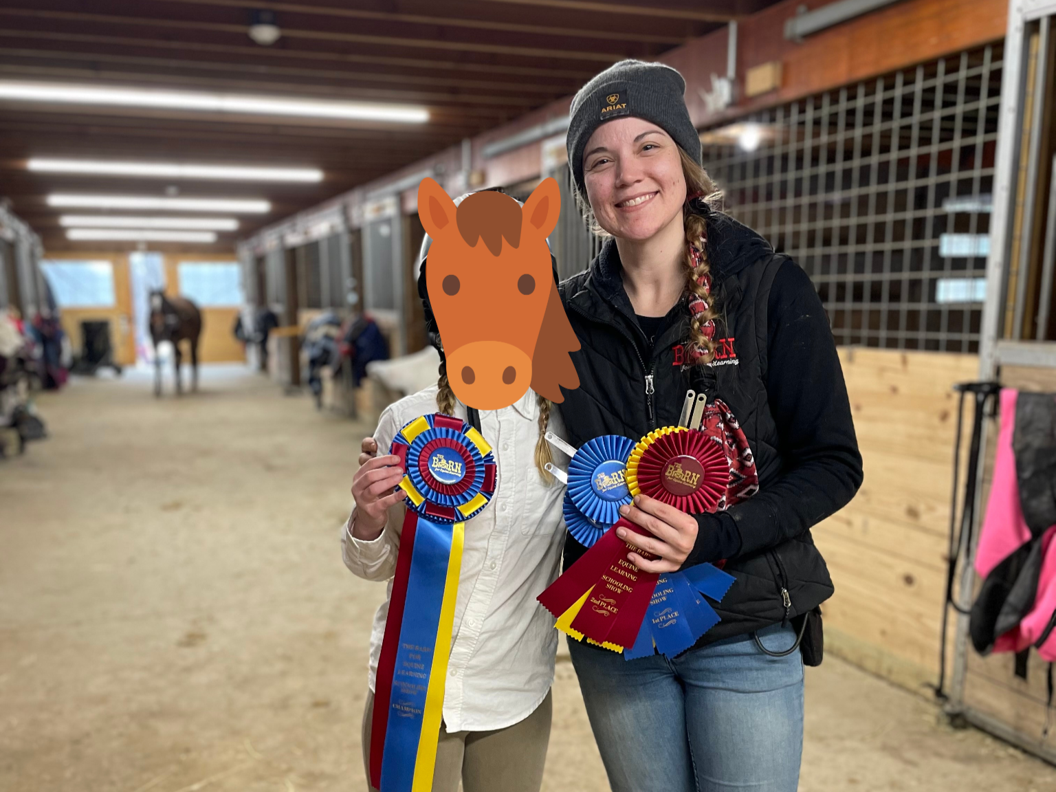 Two women standing inside a horse stable, smiling and holding award ribbons, with horses and stable stalls in the background.