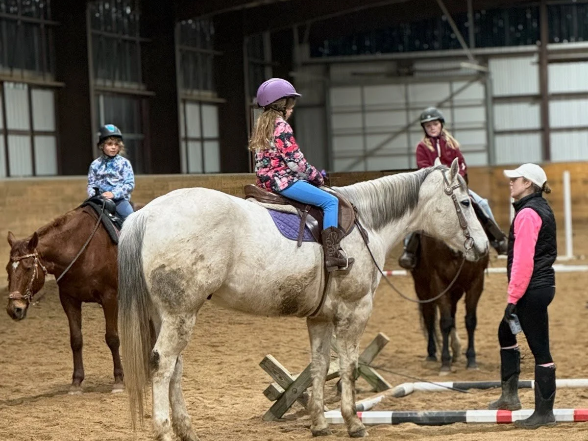 Children riding horses in an indoor arena with a trainer guiding them.