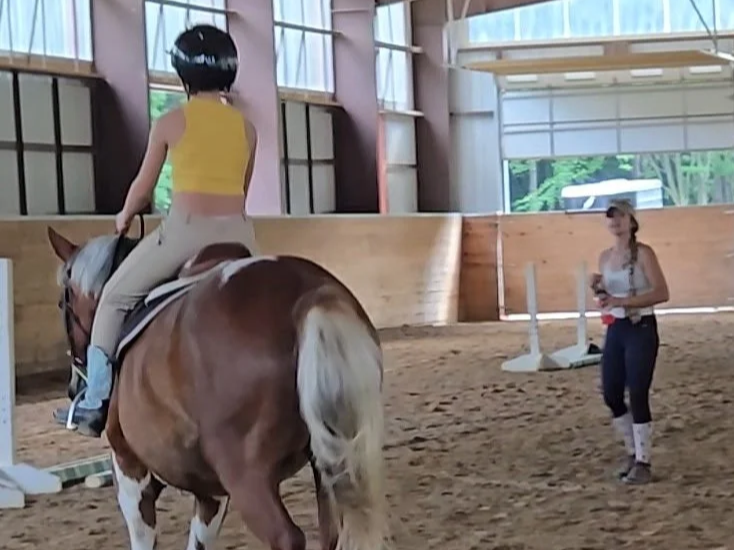 Girl riding a horse in an indoor riding arena, with a woman standing nearby.