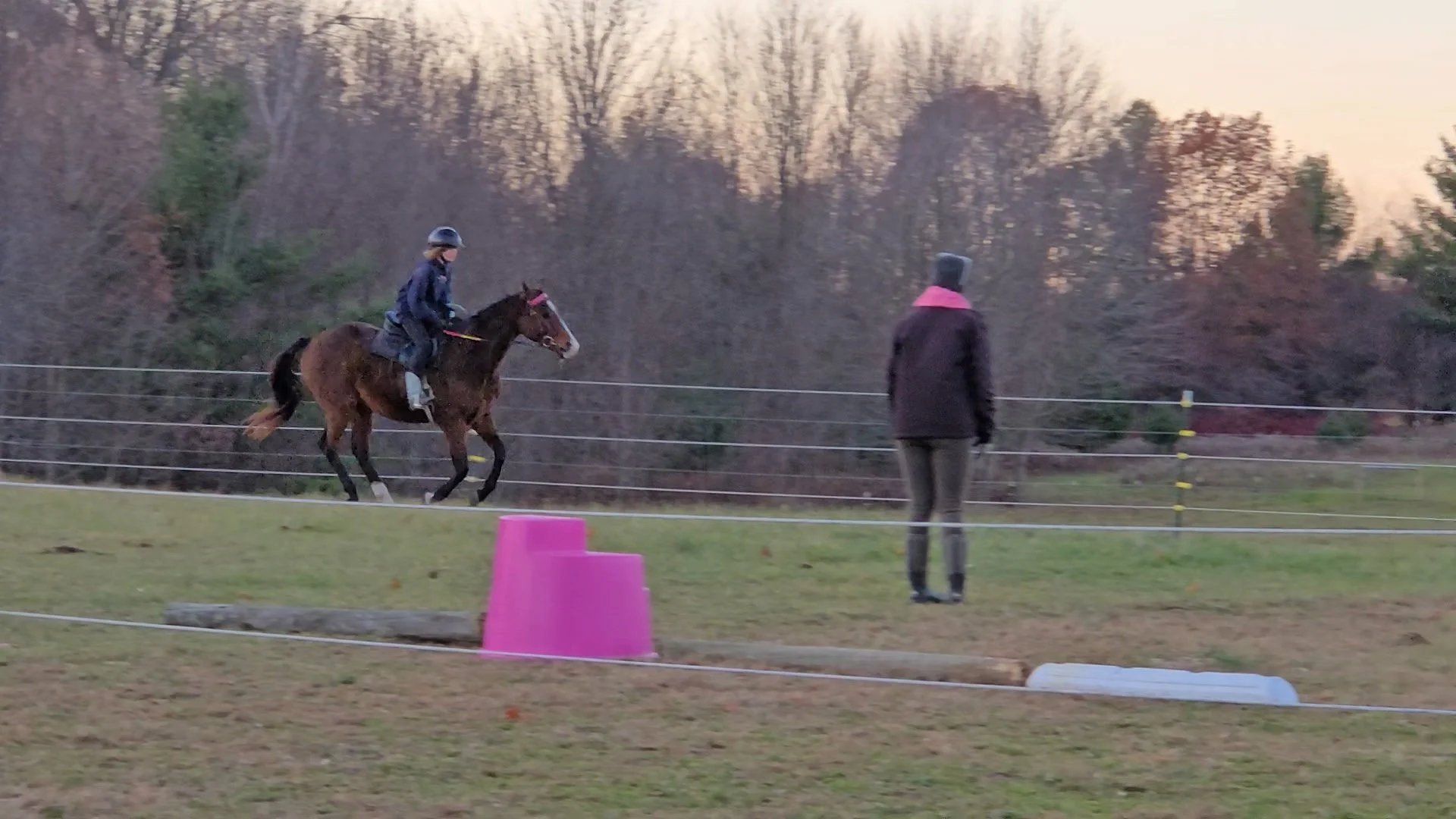 A girl riding a horse in an outdoor riding area at sunset, with an instructor or observer standing nearby. The horse is galloping, and the area is enclosed by a safety fence with pink cones and blue markers on the ground.