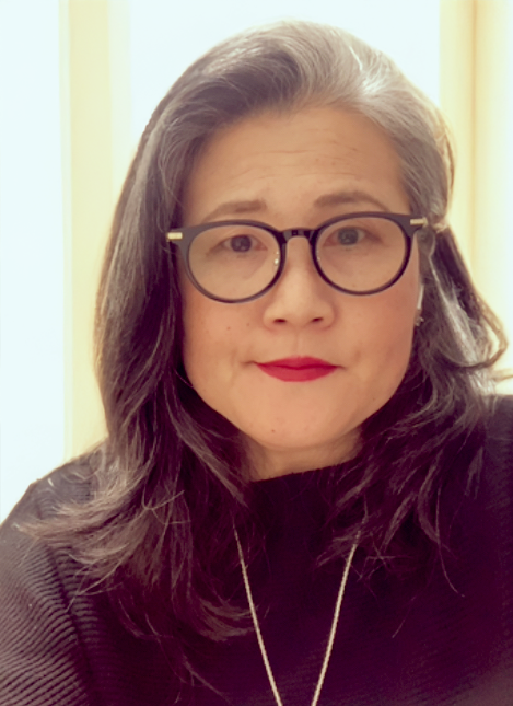 A woman with shoulder-length dark gray hair wearing black glasses with gold accents, bright red lipstick, and a black top, standing in front of a light-colored background.