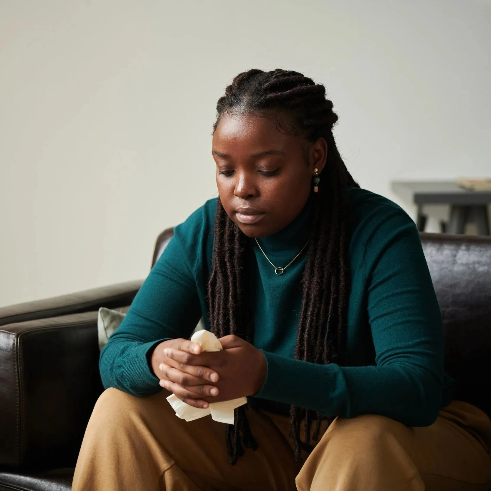 A woman with dark braided hair sitting on a brown leather couch, looking down at her hands which she is holding with a tissue or cloth, in a room with a neutral-colored wall.