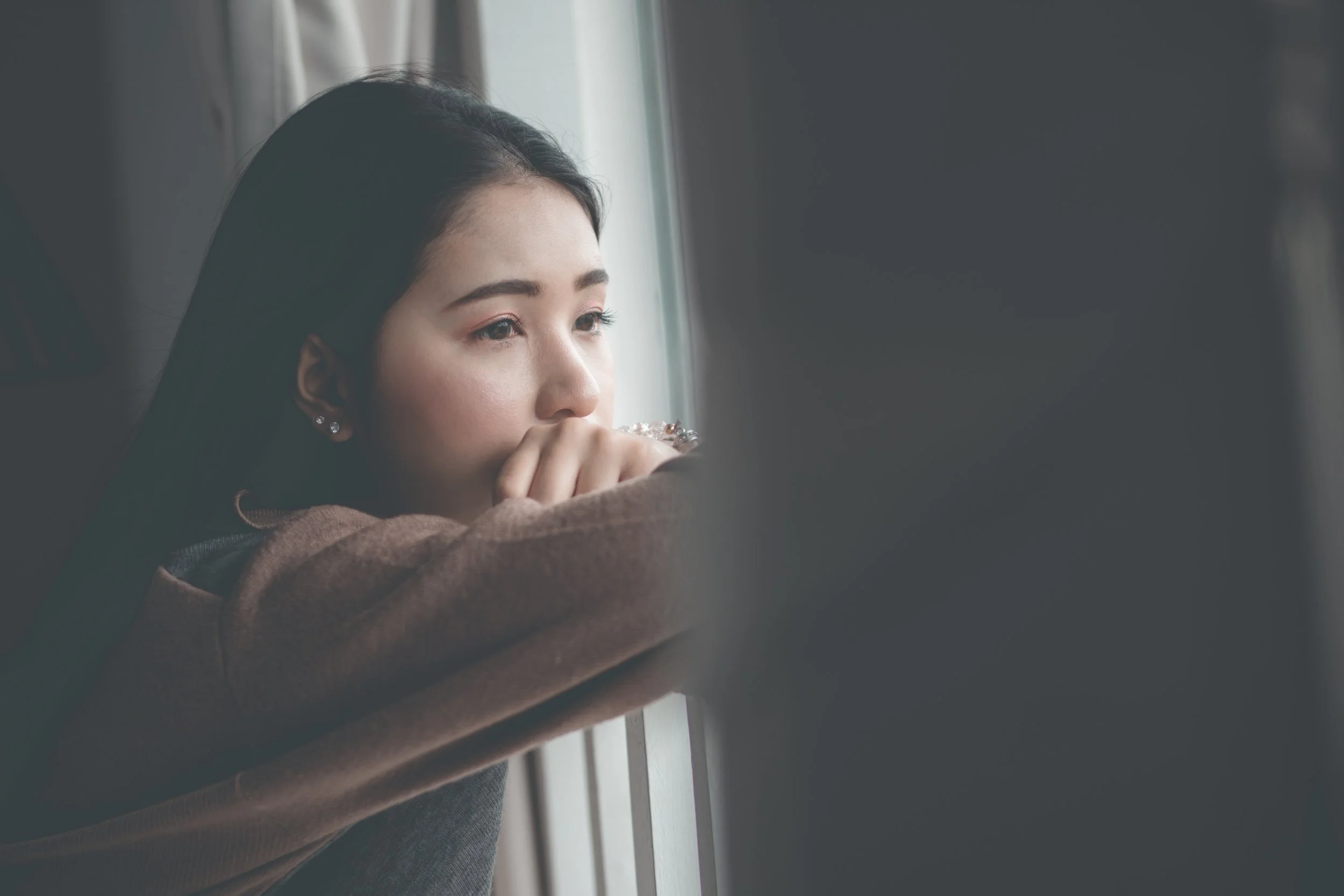 A young woman with dark hair looking thoughtfully out a window with her hand near her mouth.