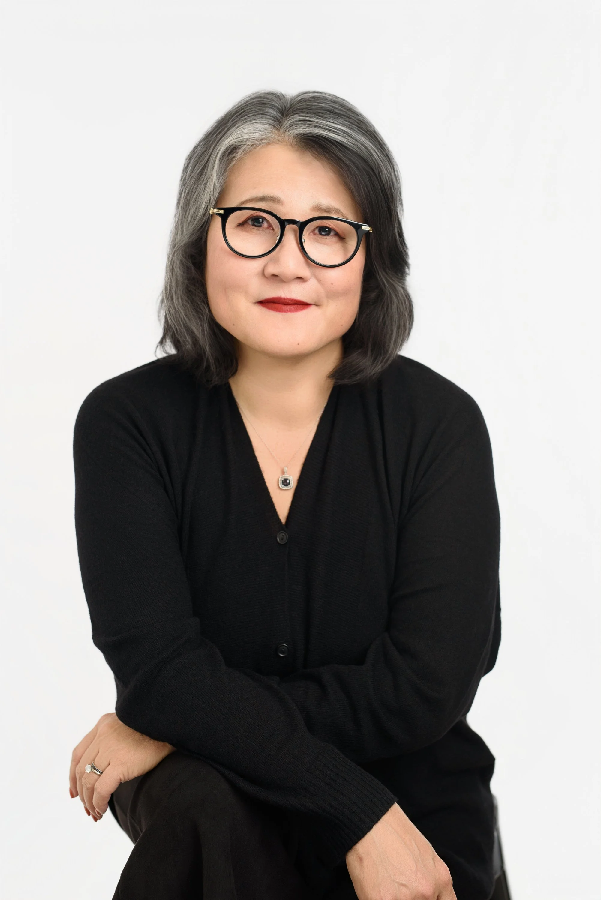 Portrait of a woman with gray hair, wearing glasses, a black sweater, and a necklace, sitting against a plain white background.