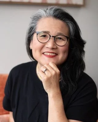 A woman with gray hair and glasses smiling, resting her chin on her hand.
