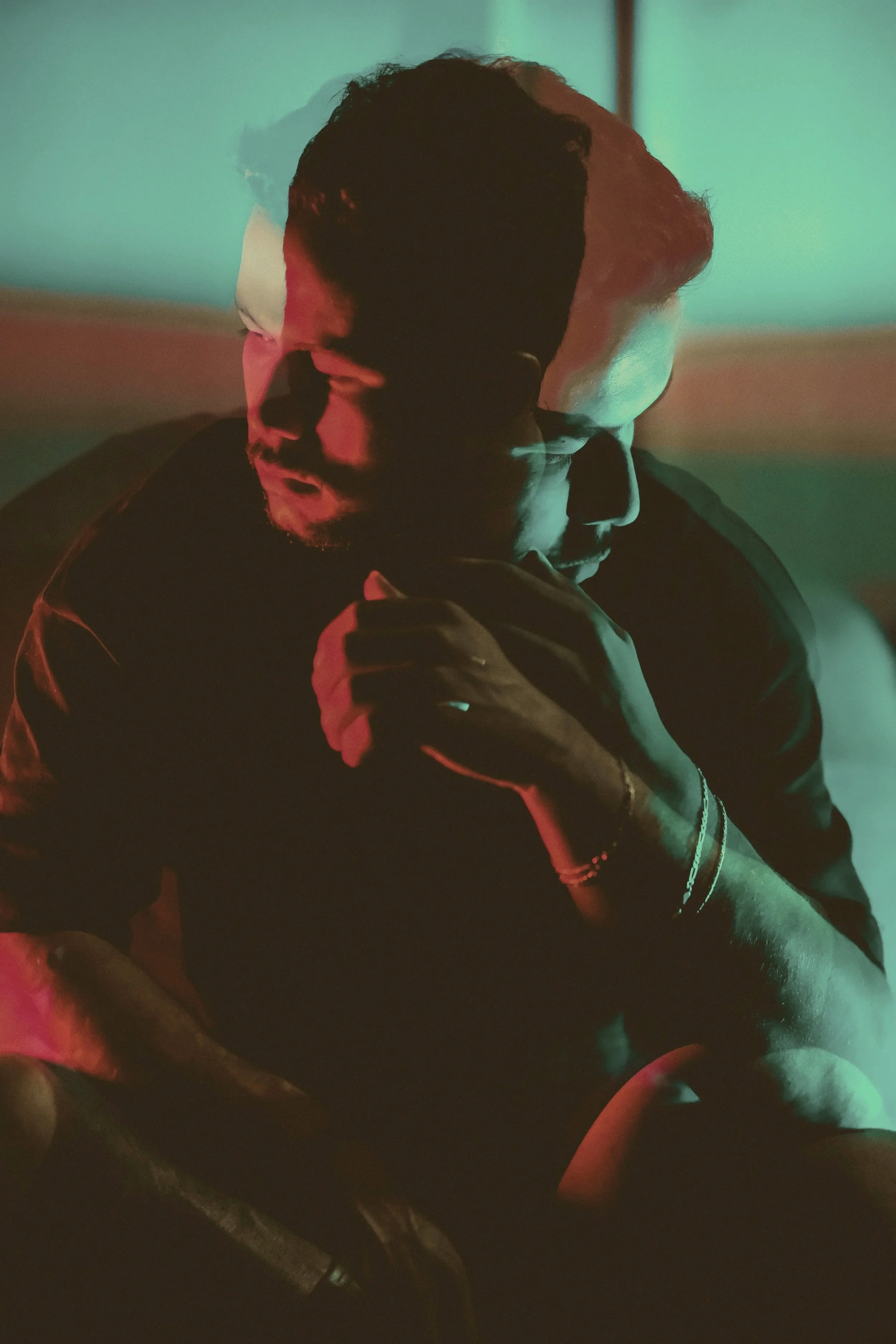 A man with a beard and glasses looking down with a hand near his face, under multicolored lighting with red and blue hues.