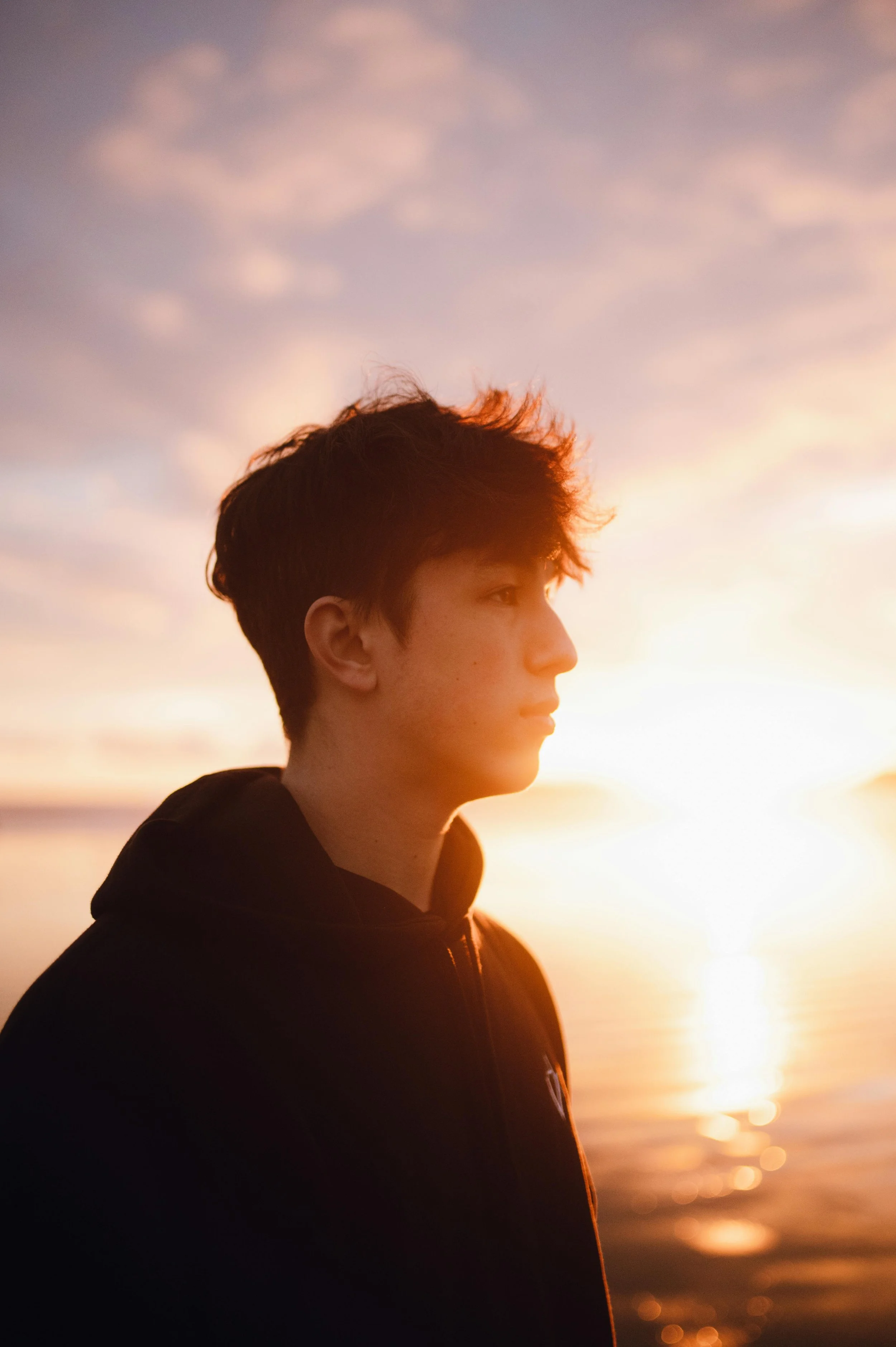 Side profile of a young man with dark hair wearing a black hoodie, standing outdoors during sunset, with the sky filled with soft clouds and the sun near the horizon creating a warm glow.