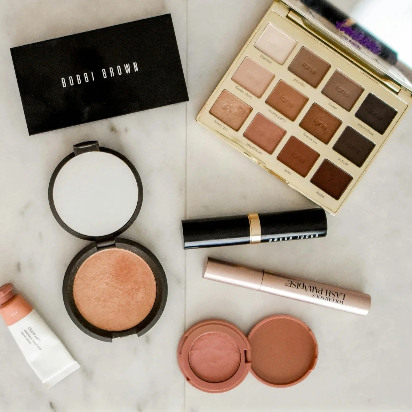 Collection of makeup products including an eyeshadow palette, highlighter, blush, lipstick, lip gloss, and a compact mirror on a marble surface.