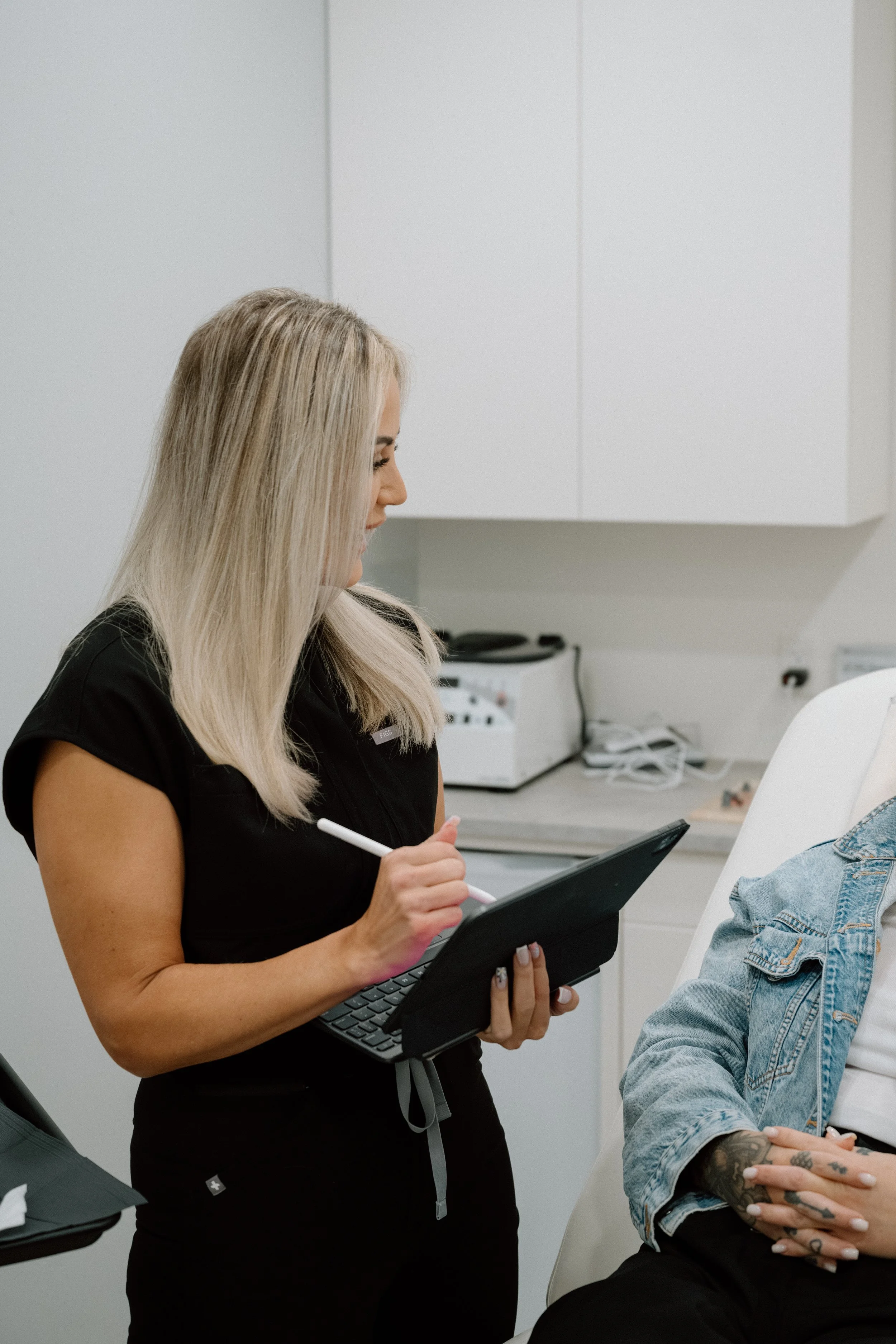 A nurse standing beside clinic chair, speaking with patient, holding a chart and pen in her hands, smiling with blonde hair and black scrubs