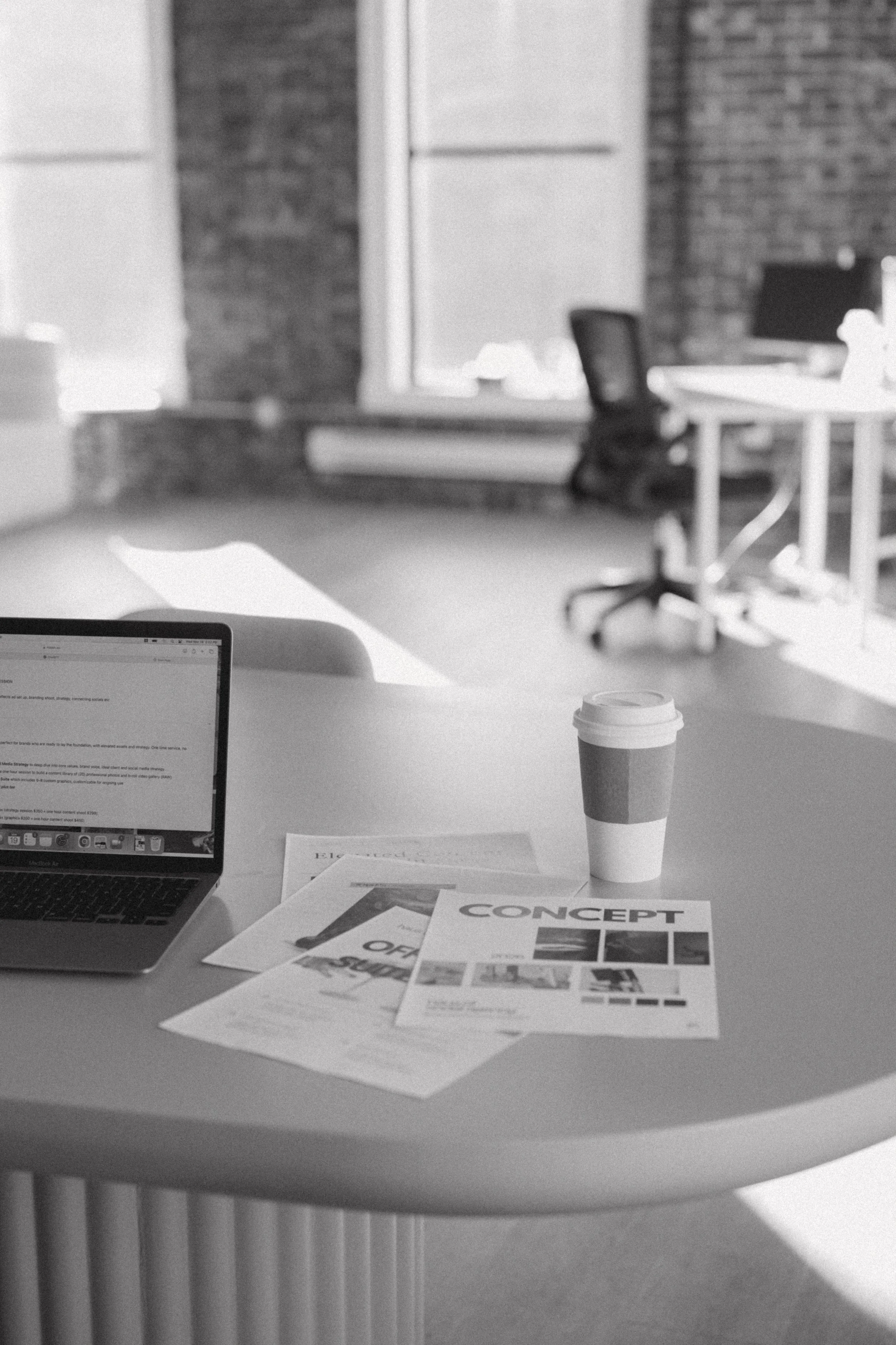 Black and white photo of a work desk with a laptop, a paper coffee cup, and some printed documents labeled "CONCEPT." In the background, there are office chairs and large windows letting in natural light.
