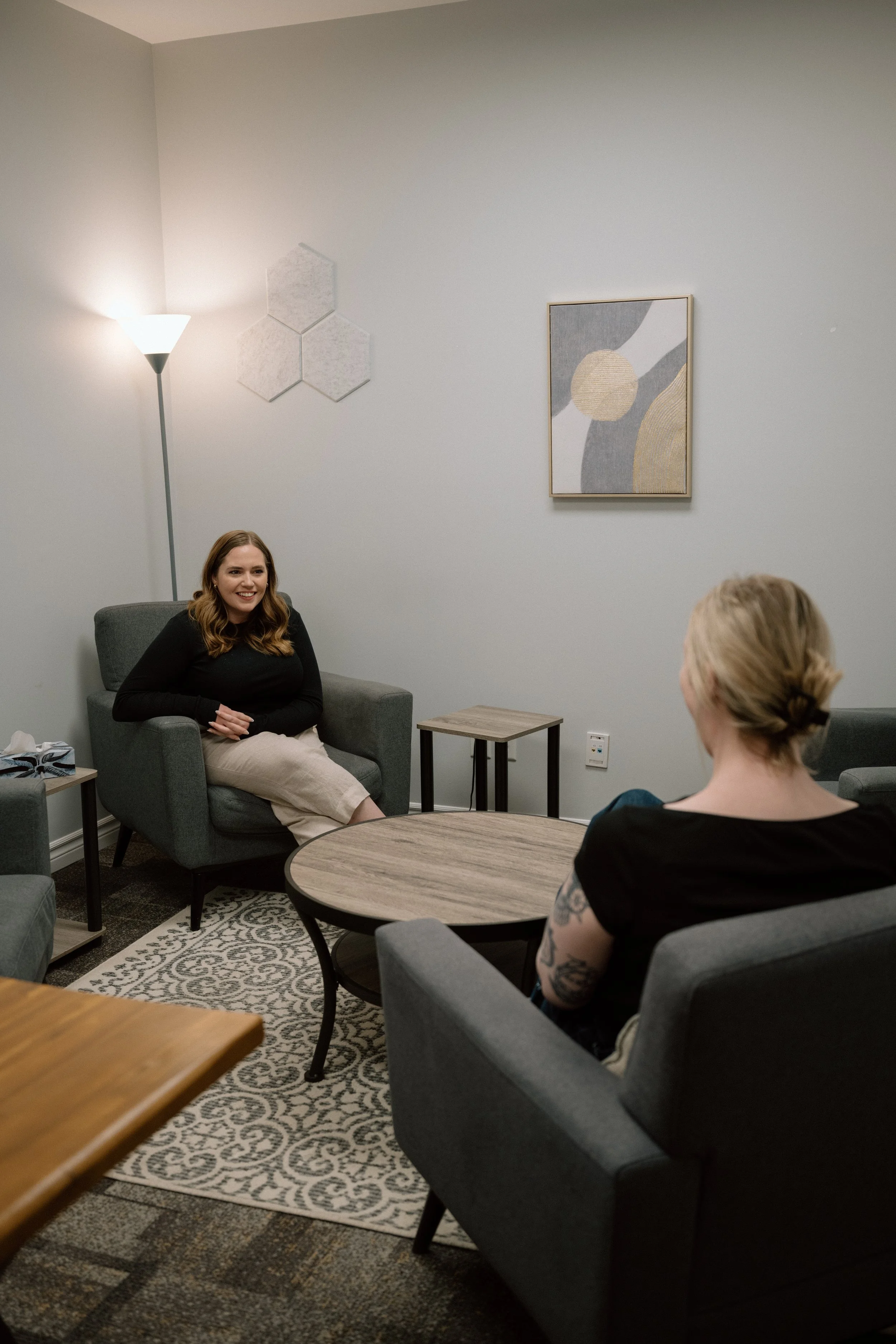 Two females sitting in a therapy room, engaged in conversation, muted tones and grey furniture during therapy session