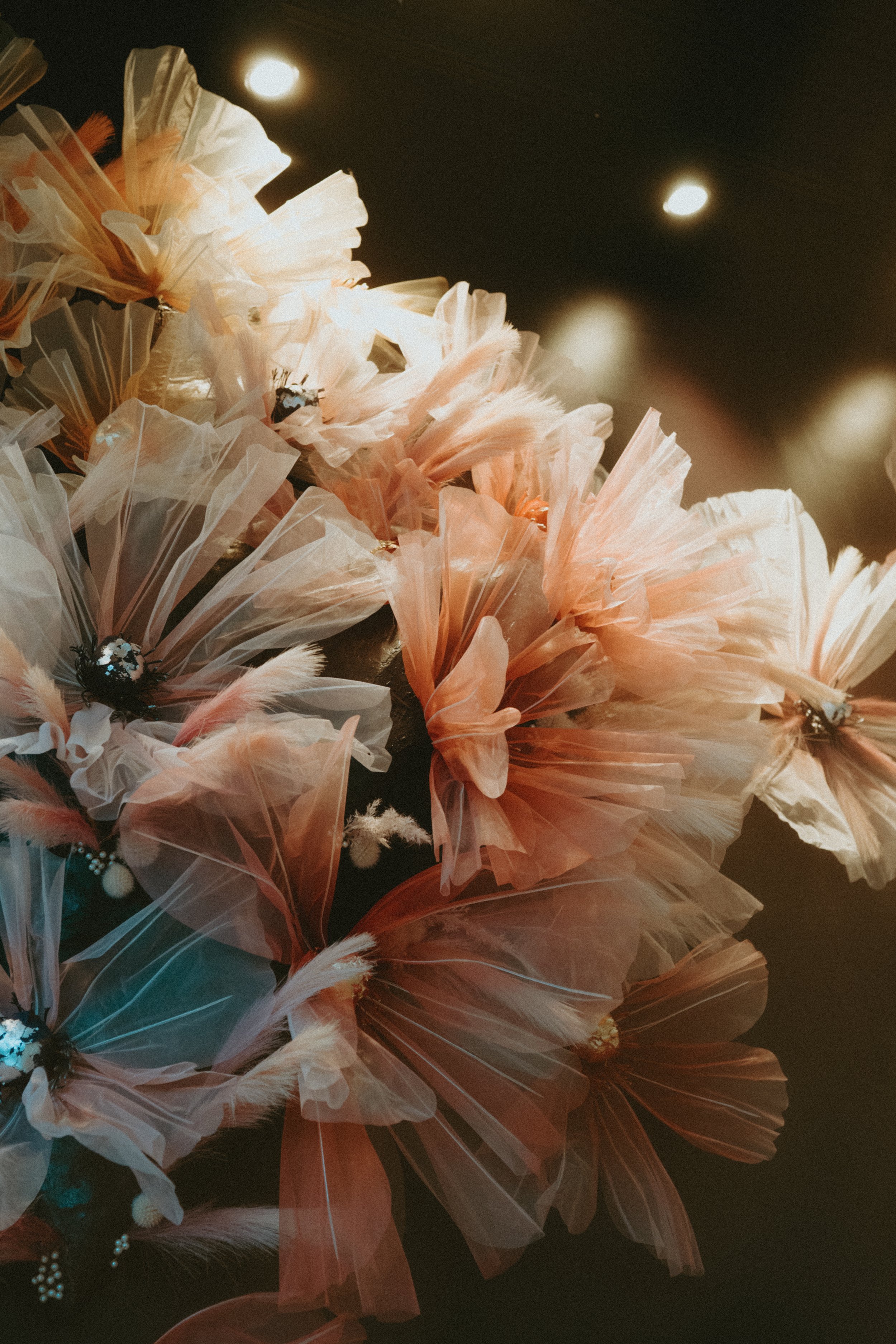 Artificial flowers made of pink and white tissue paper, arranged on a dark surface with overhead lighting.