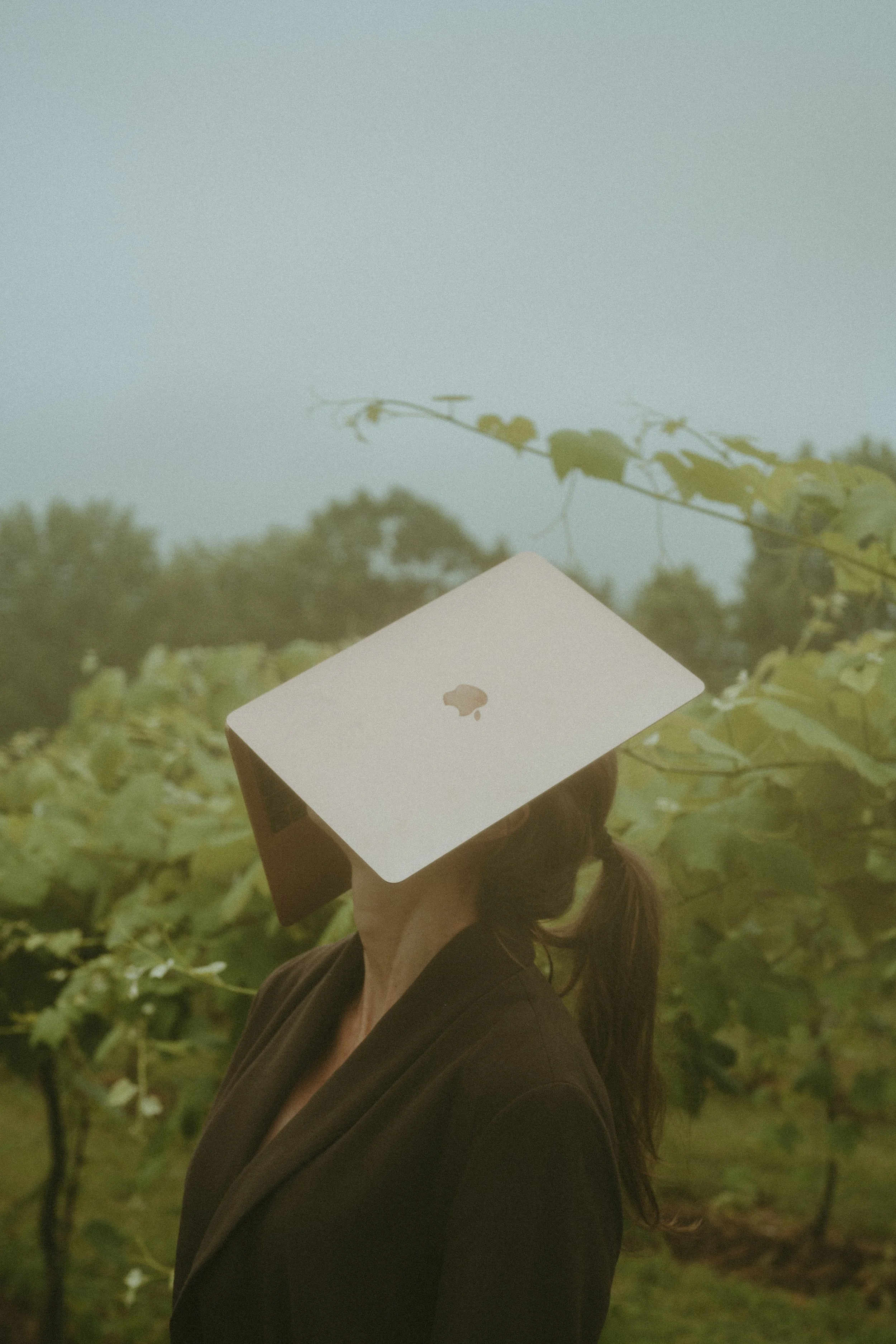 A person outdoors with a MacBook laptop with the Apple logo on their face, against a background of greenery and trees.