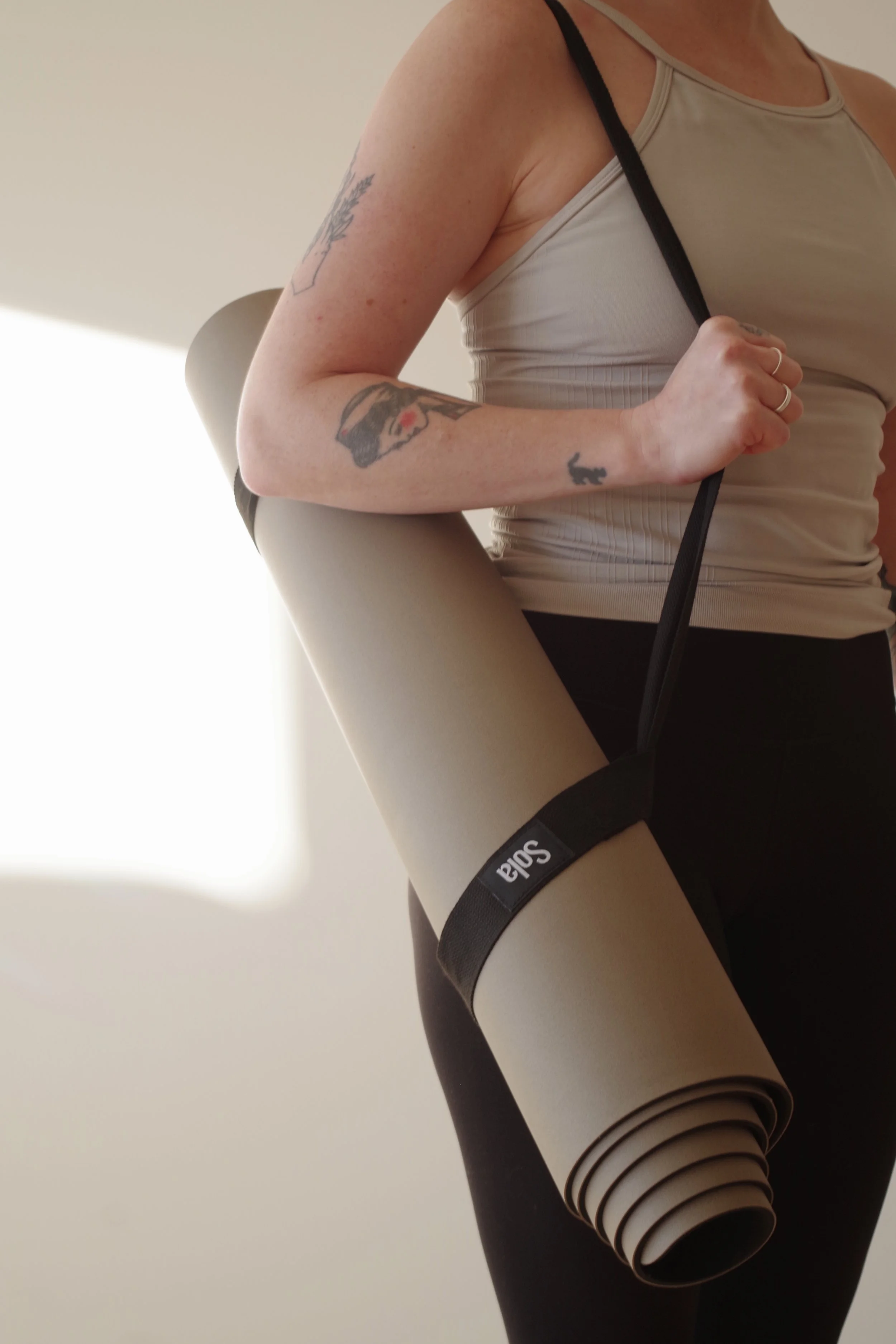 A female with a yoga mat in a carrying strap around her shoulder, beige yoga mat labelled "Sola", with a beige tank top, black pants and a plain background with sunlight