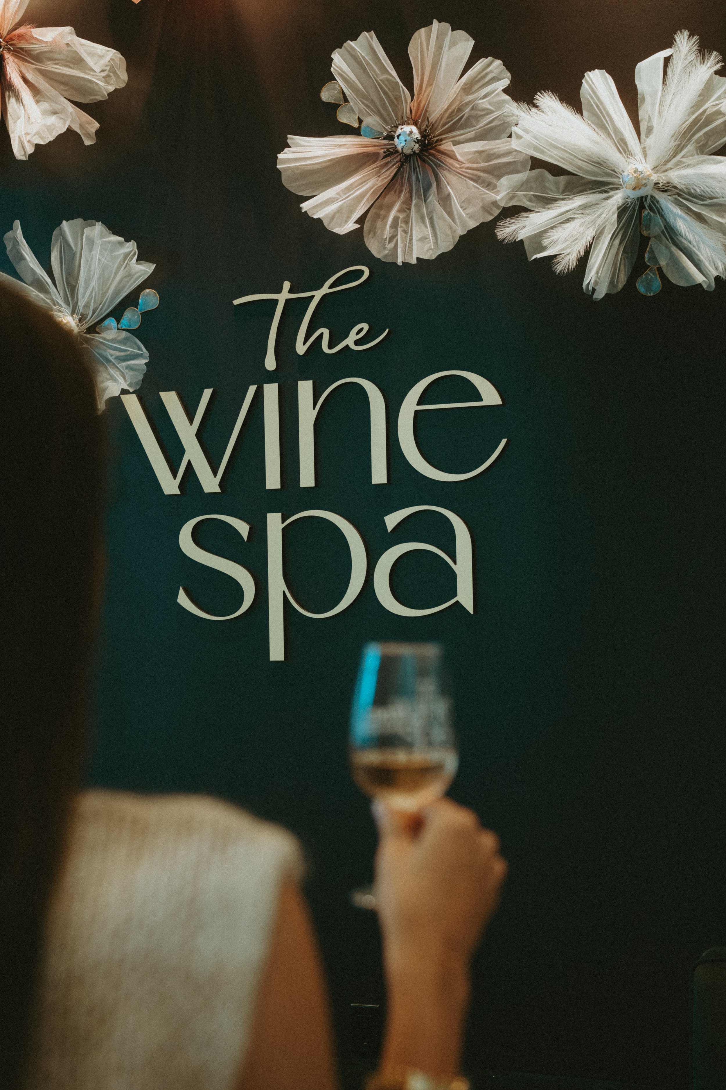 Person holding a glass of rosé wine in front of a dark wall with white floral decorations and the words 'The wine spa' written on it.