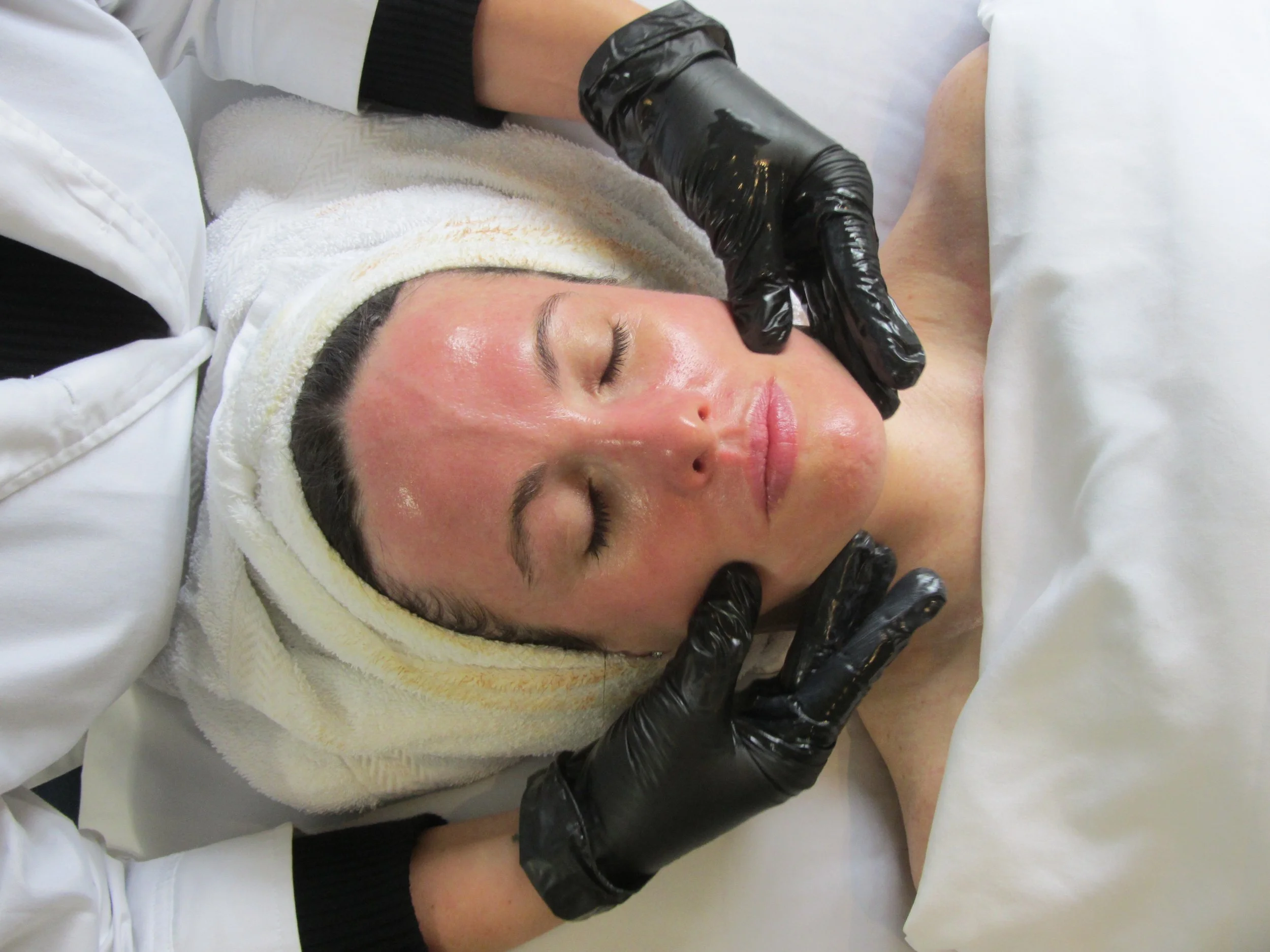 A birds-eye-view photo of a female, lying down with her eyes closed during a facial. Glowing skin, a towel wrapped around her hair. Two hands with black gloves appear to be giving facial massage 