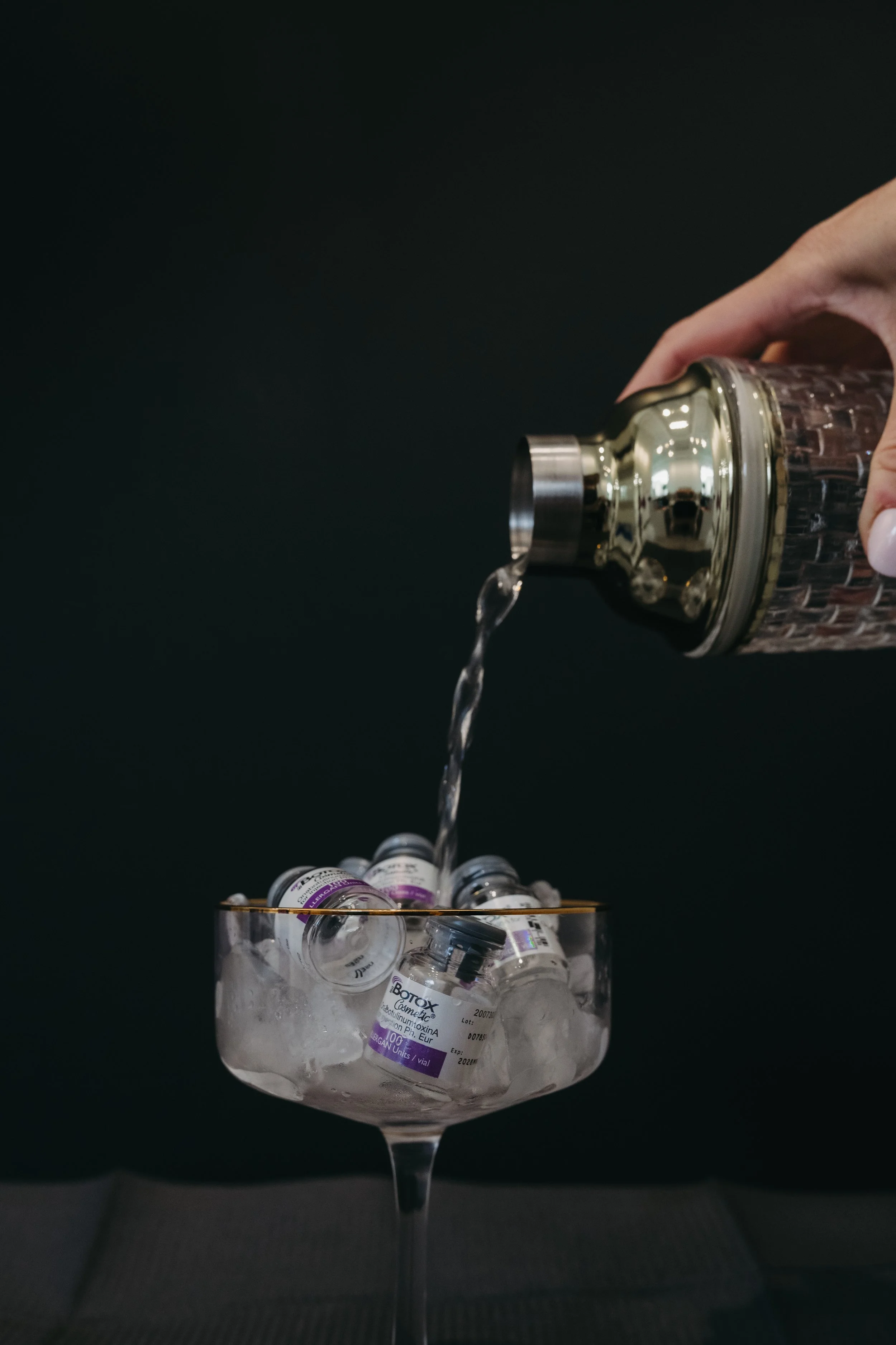 a martini glass full of Botox (neuromodulator) vials, with liquid being poured in from a cocktail shaker, with a black background