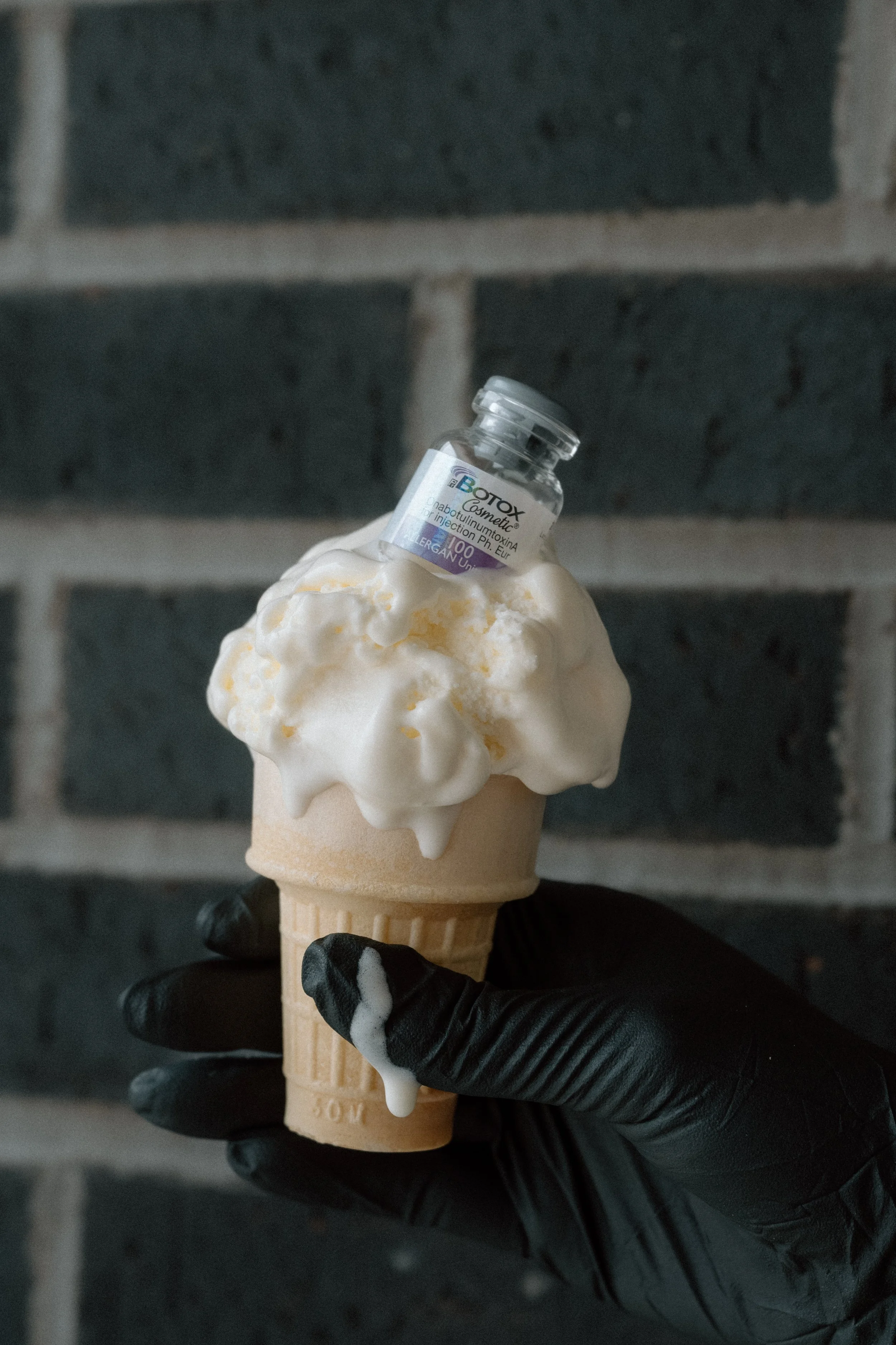 A bottle of neuromodulator (Botox) sitting on top of an ice cream cone, with melting vanilla ice cream, held against a dark brick background