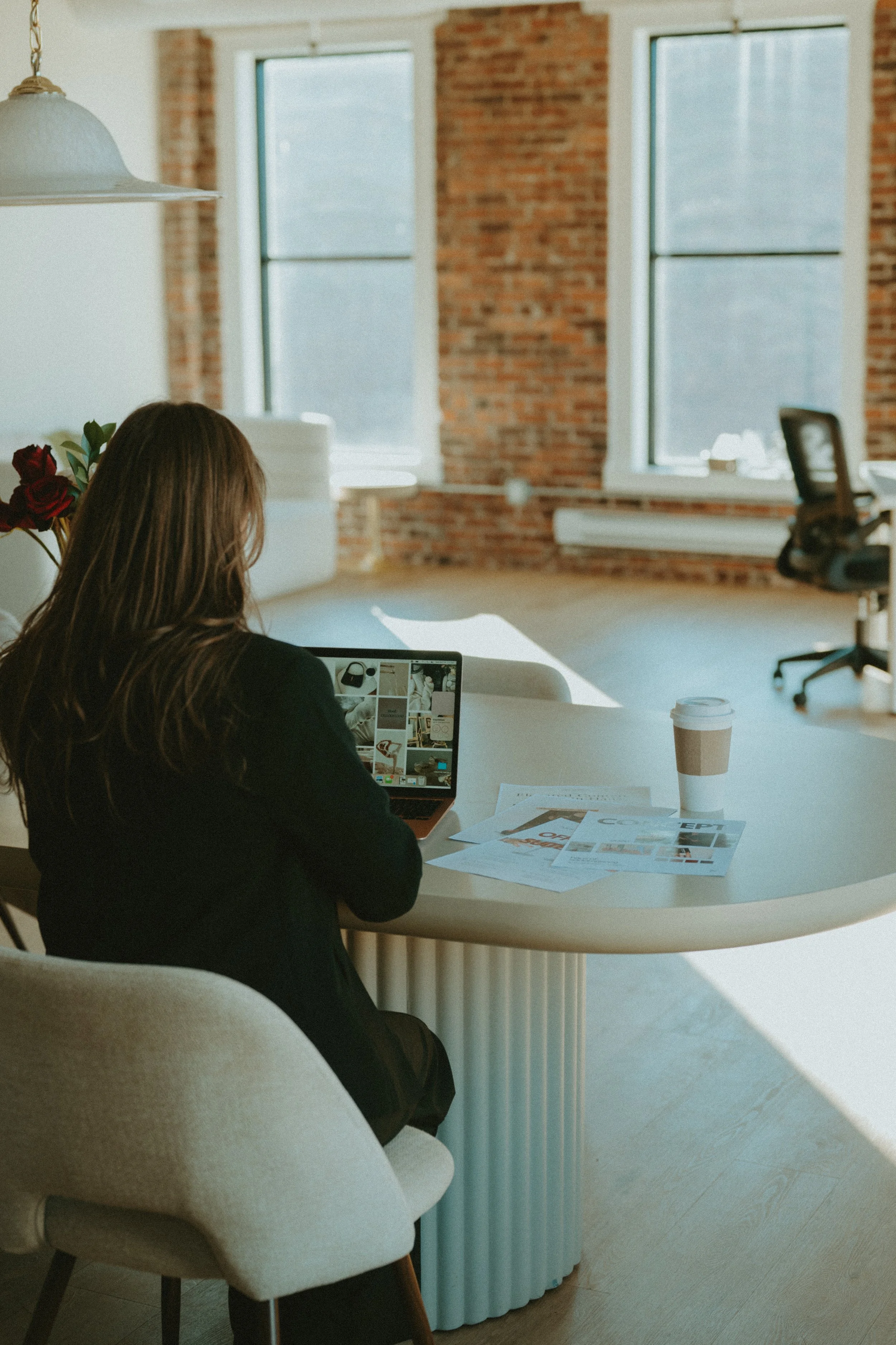 Woman working at a desk in a bright, industrial-style office with brick walls and large windows, using a laptop with magazines and a coffee cup on the table.