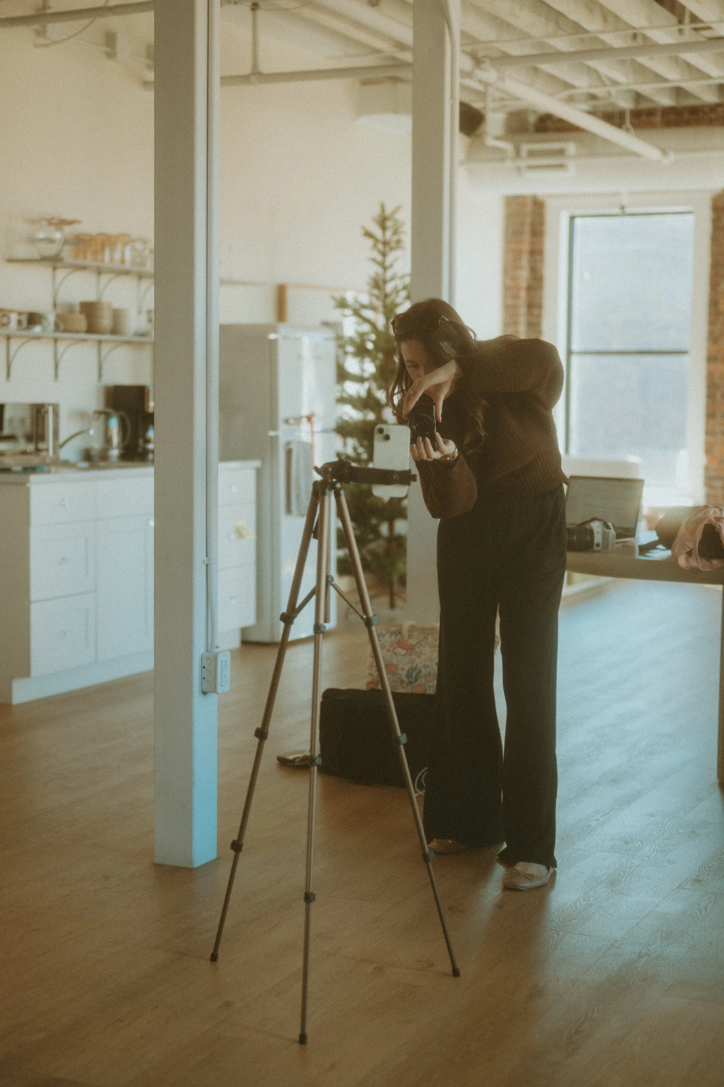 A woman is taking a photo with a camera on a tripod inside a room with large windows and a Christmas tree in the background.