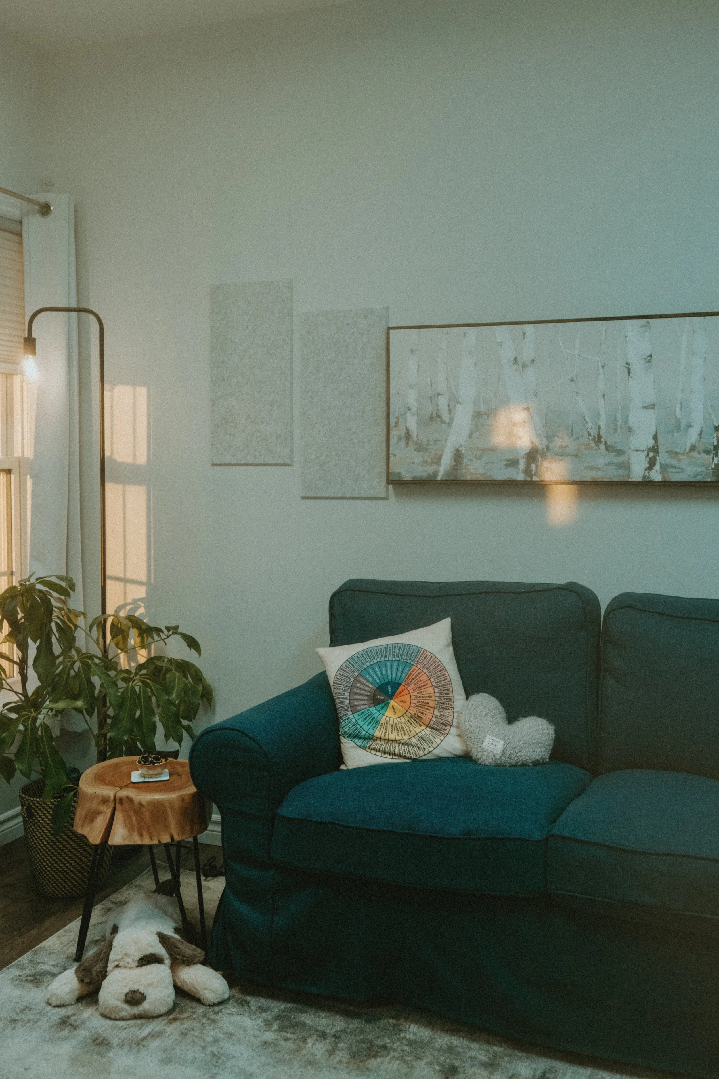Living room with a dark teal sofa, decorative pillows, a small wooden side table, a lamp, a potted plant, a white shaggy dog plush, artwork on the wall, and sunlight streaming through a window.