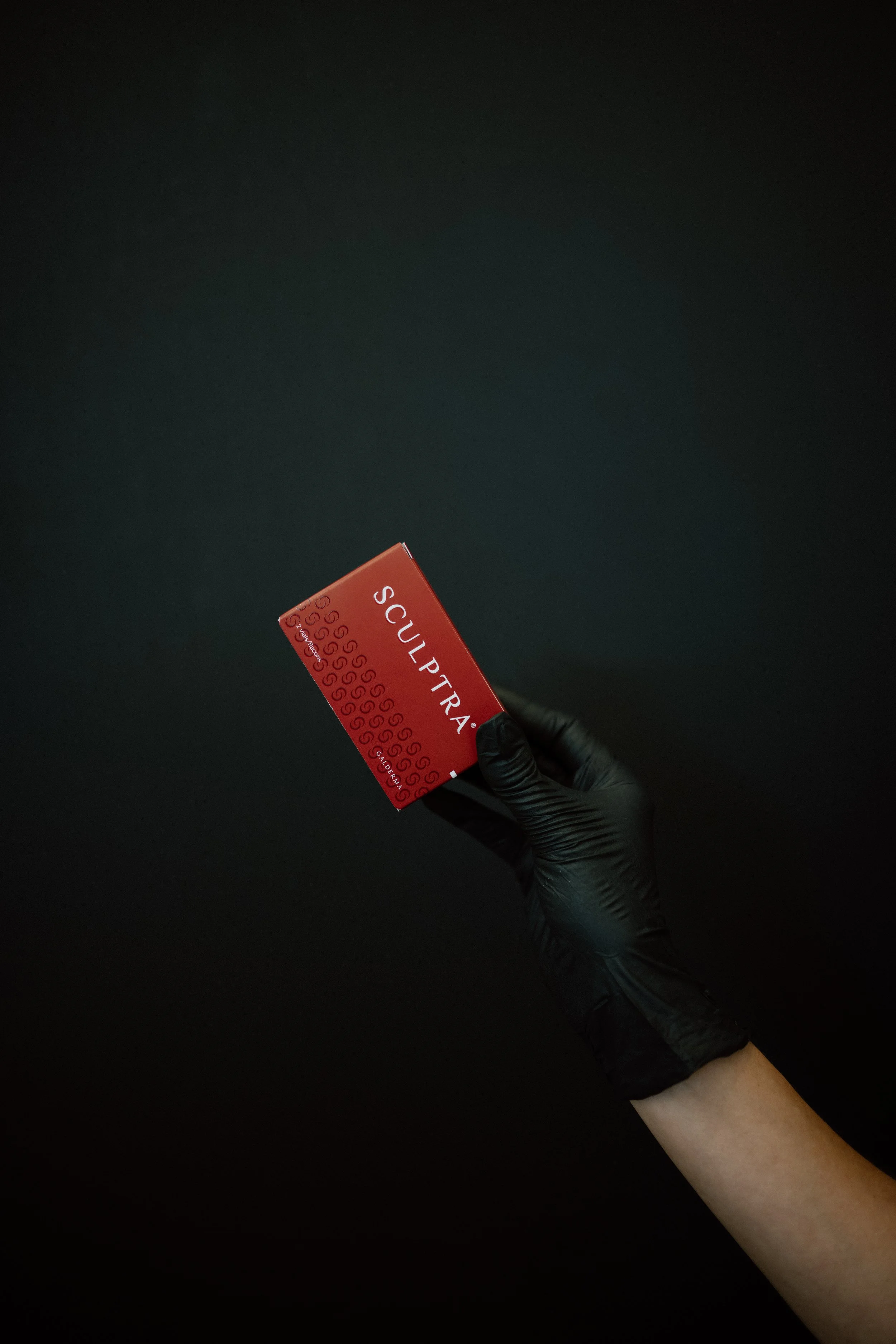 A black-gloved hand, holding a red product box for Sculptra, against a black background