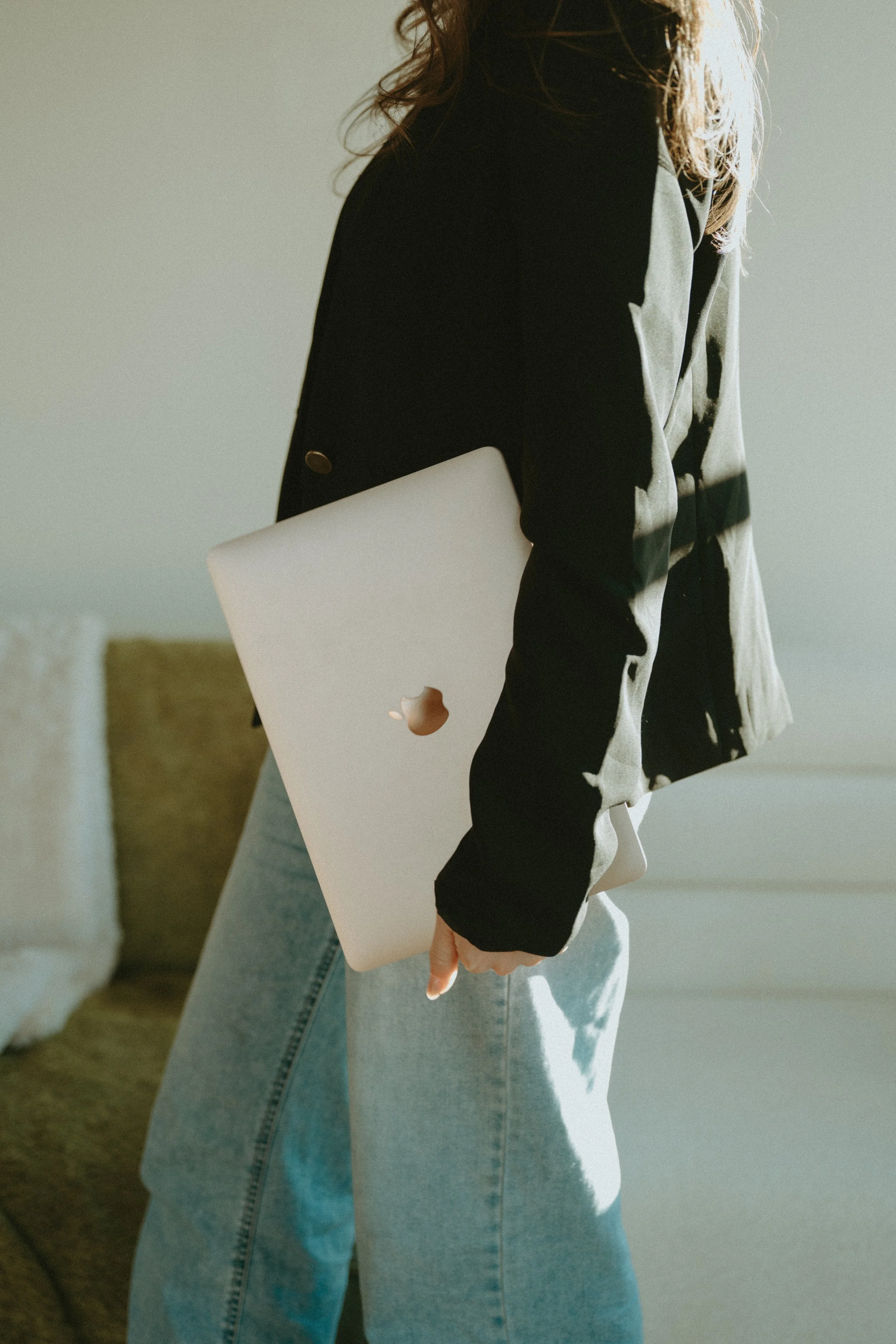 Person in a black jacket holding a closed silver MacBook in their right hand, standing indoors.