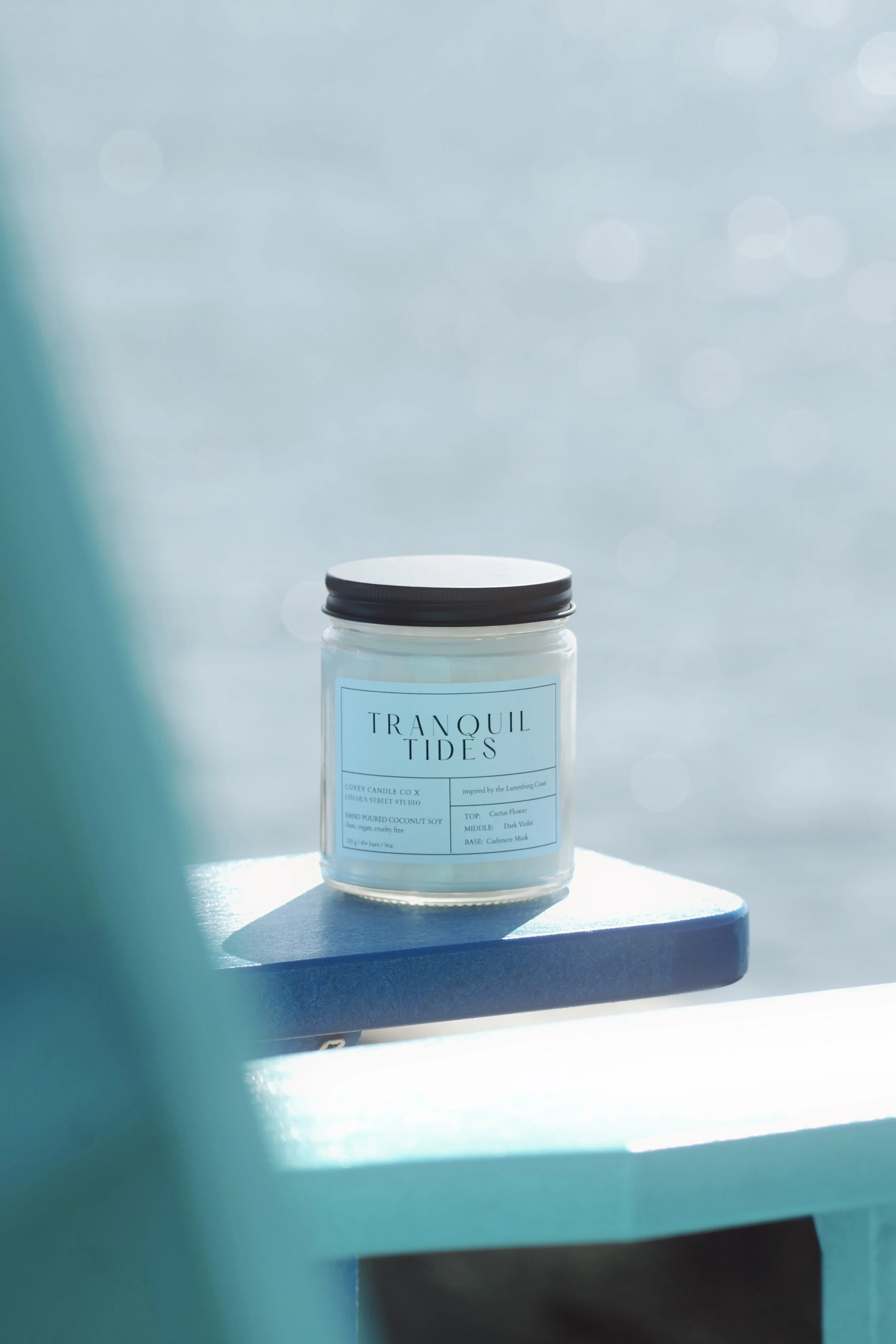 A white candle in a glass jar with a black lid, labeled 'Tranquil Tides,' sits on a blue surface with a blurred background.