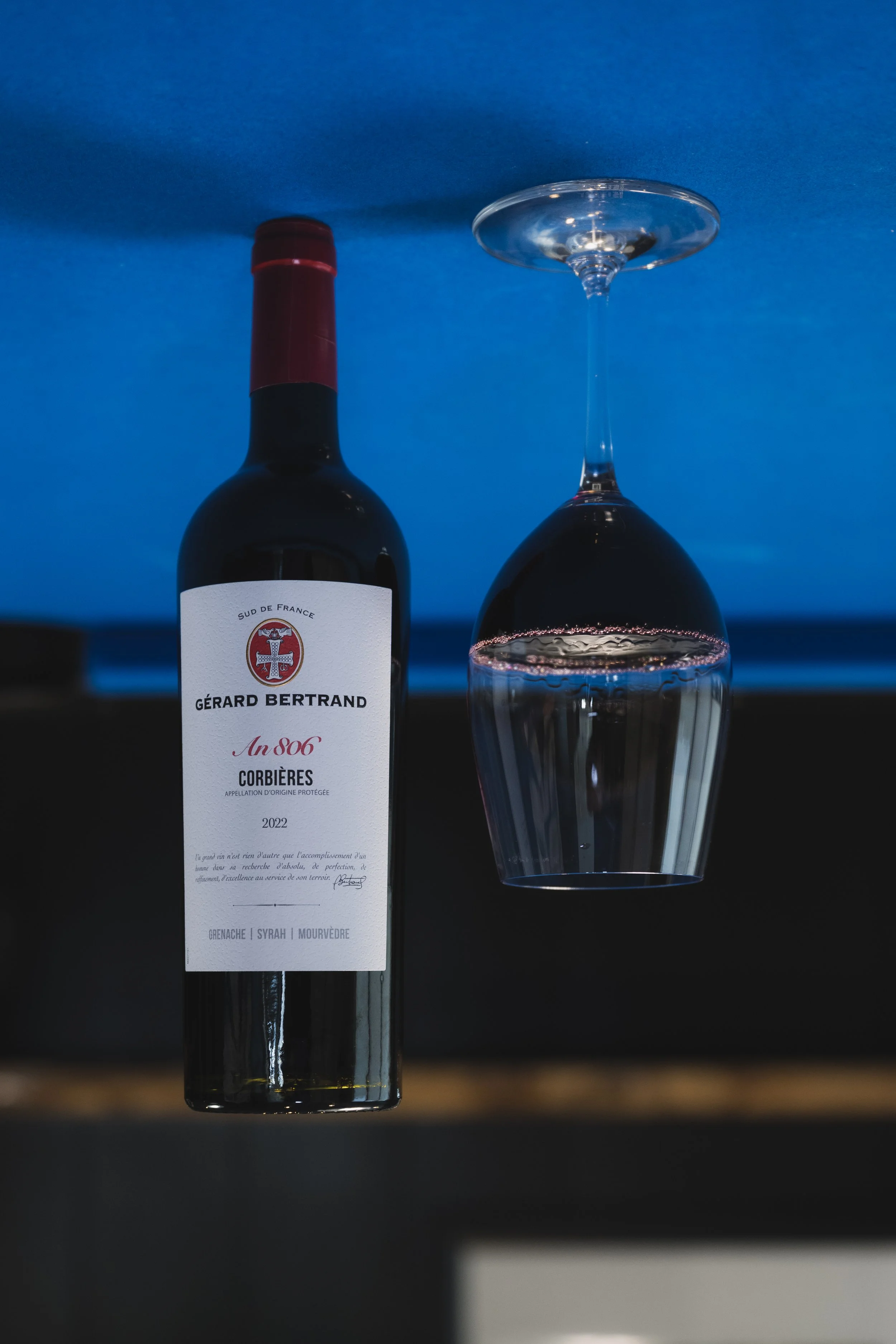 A red wine bottle, perched upside down on a blue background, next to a half-full wine glass, also upside down