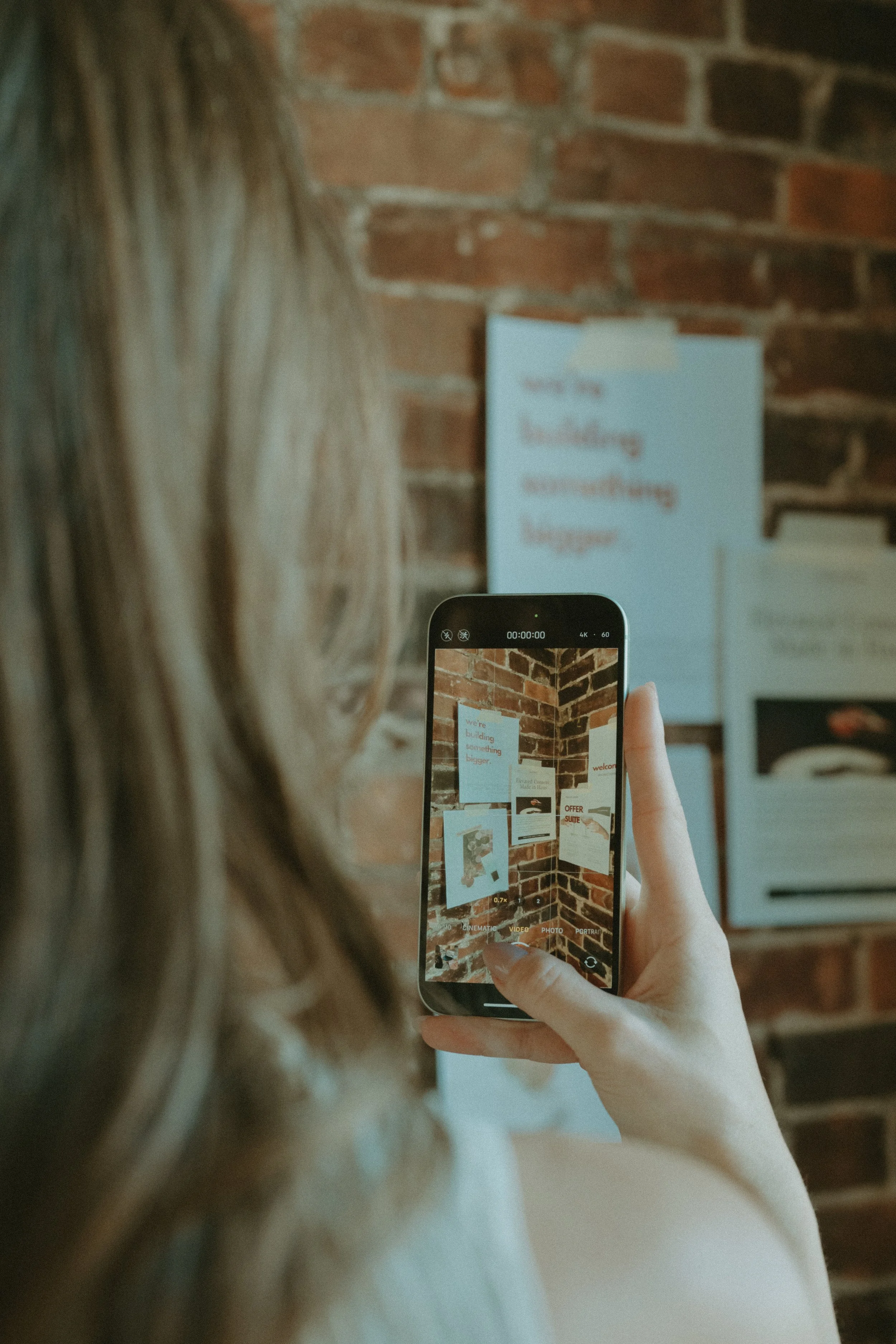 A woman with light brown hair taking a photo with her smartphone, capturing a display of posters or signs on a brick wall.