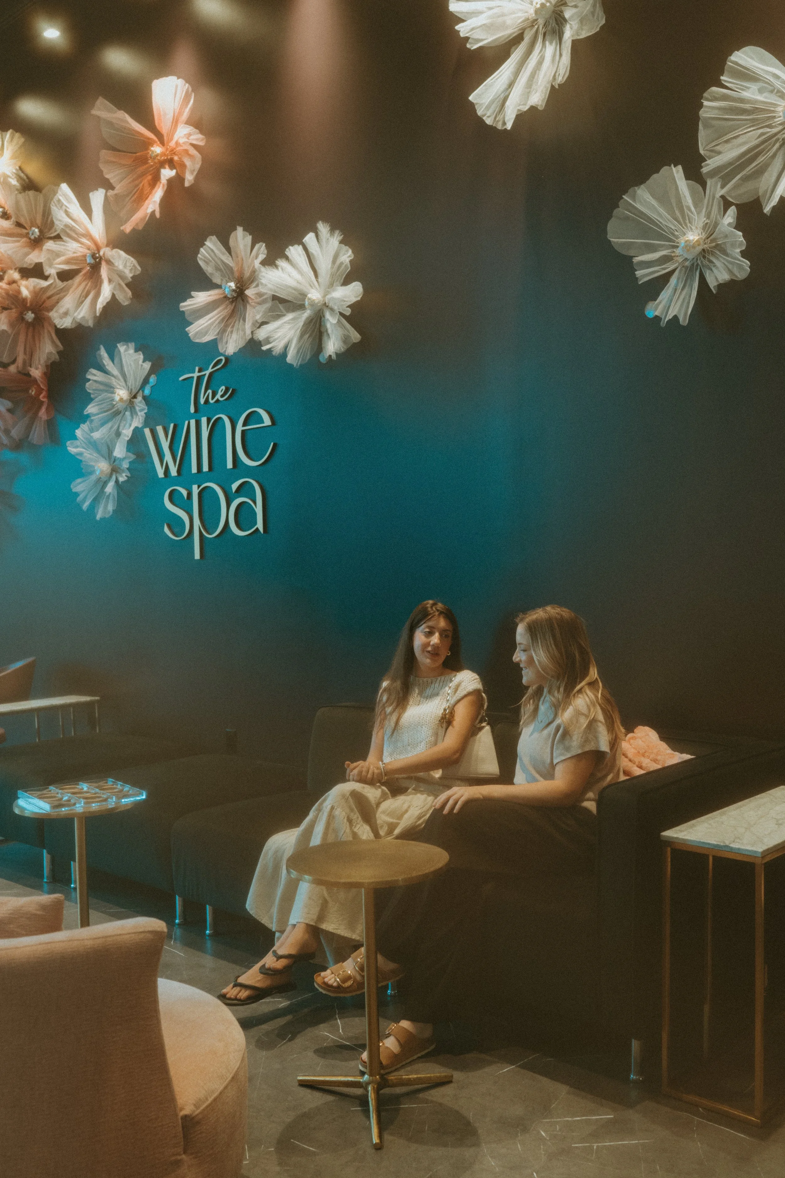 Two women sitting on a black sofa in a wine spa, engaged in conversation, with decorative white flowers on the wall behind them.