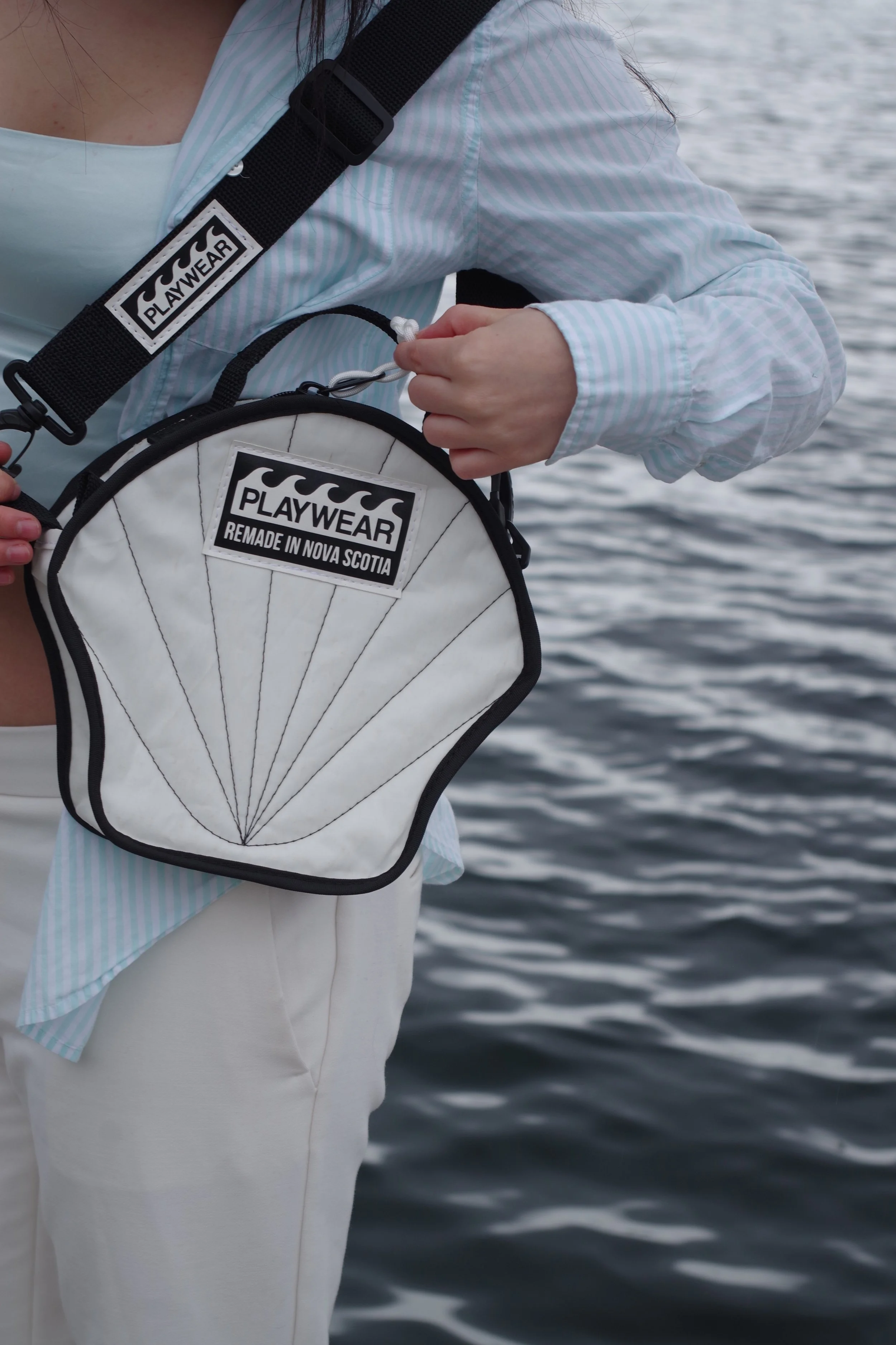 Person wearing light-colored pants and a light blue and white striped shirt, carrying a white bag with black trim and a 'PLAYWEAR' label, standing near water.