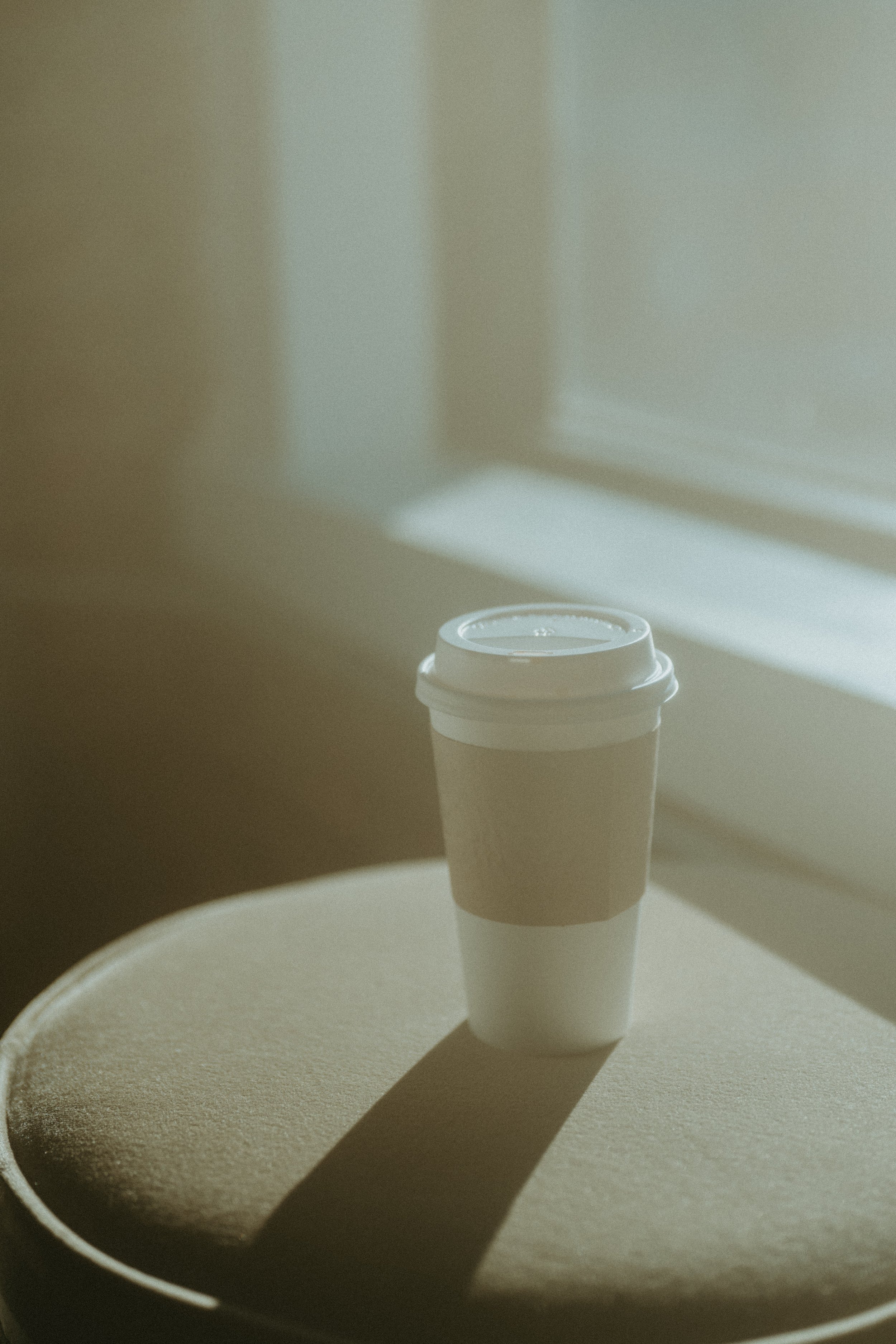 A white disposable coffee cup with a plastic lid sitting on a beige circular table near a window, casting a shadow.