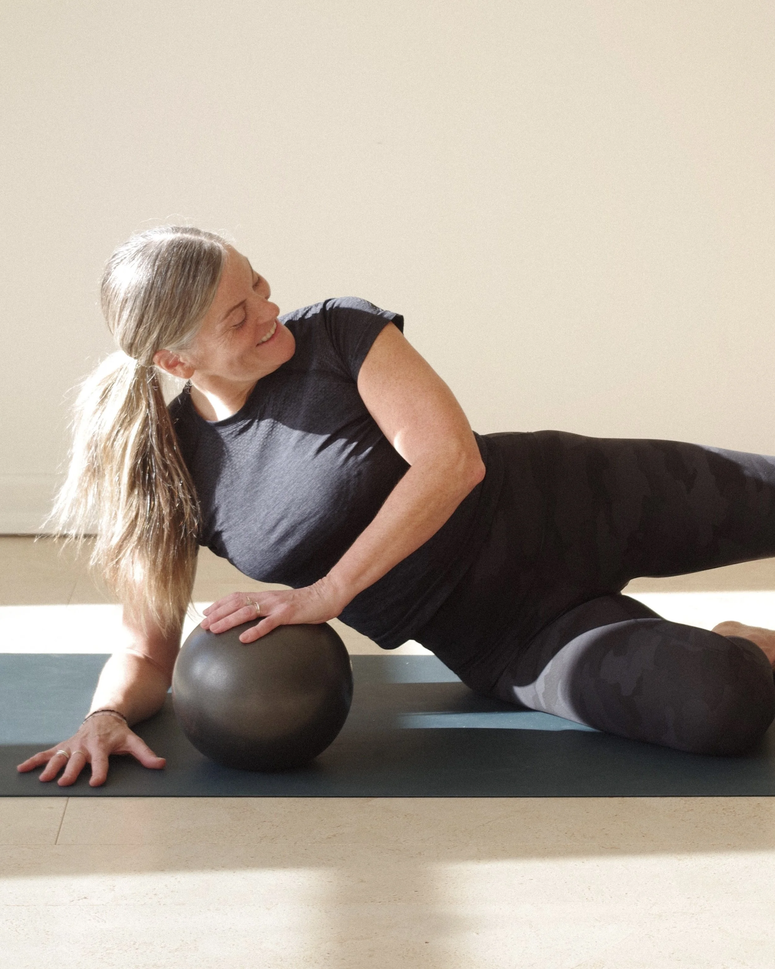 Woman performing a side exercise with a black exercise ball on a yoga mat in a bright room.