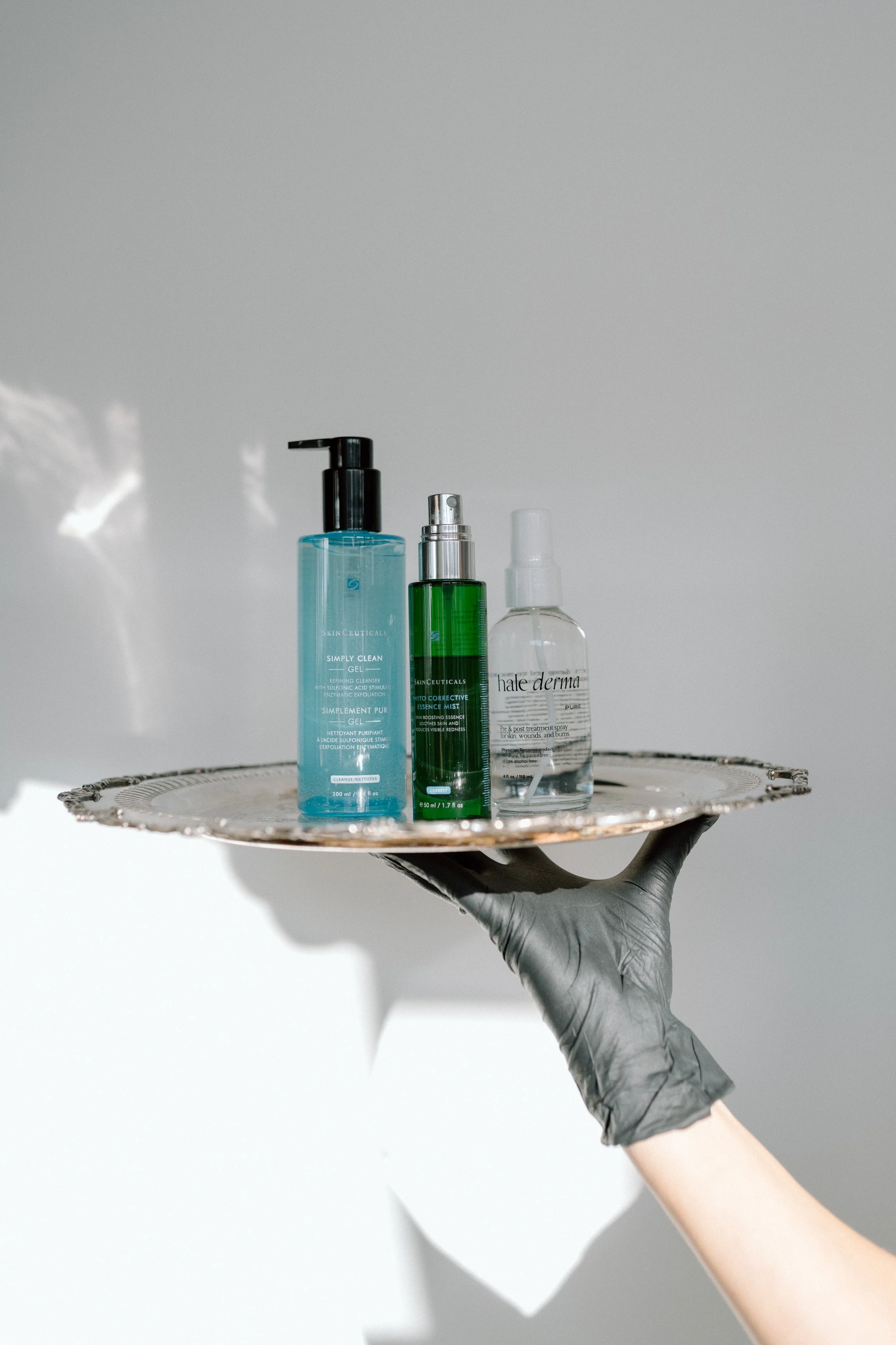 A black-gloved hand, holding up a silver tray, that has multiple skincare products on it in the sunlight, against a white plain wall