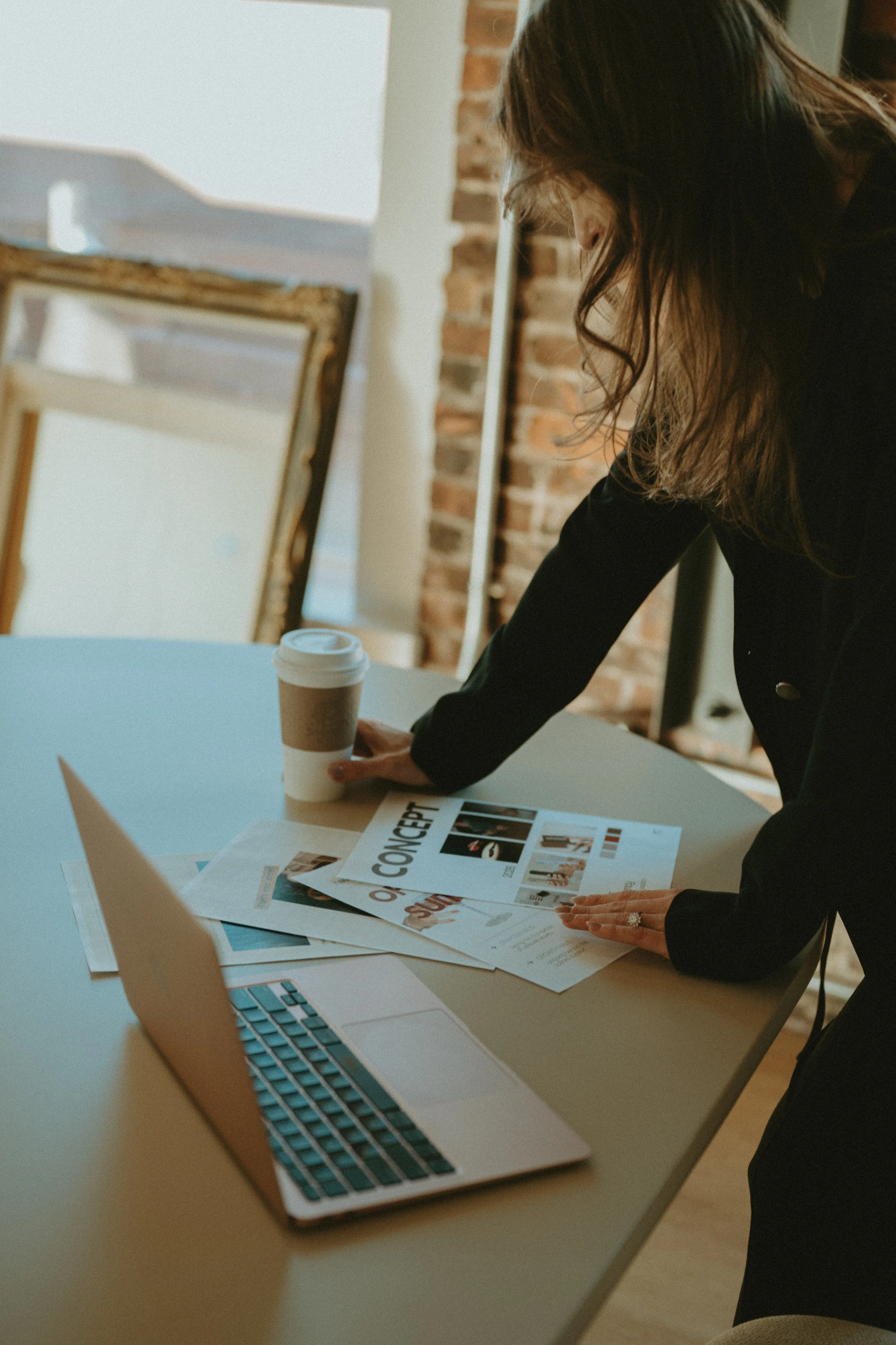 A woman organizing papers and a laptop on a table, with a coffee cup nearby in a modern office space with exposed brick wall.