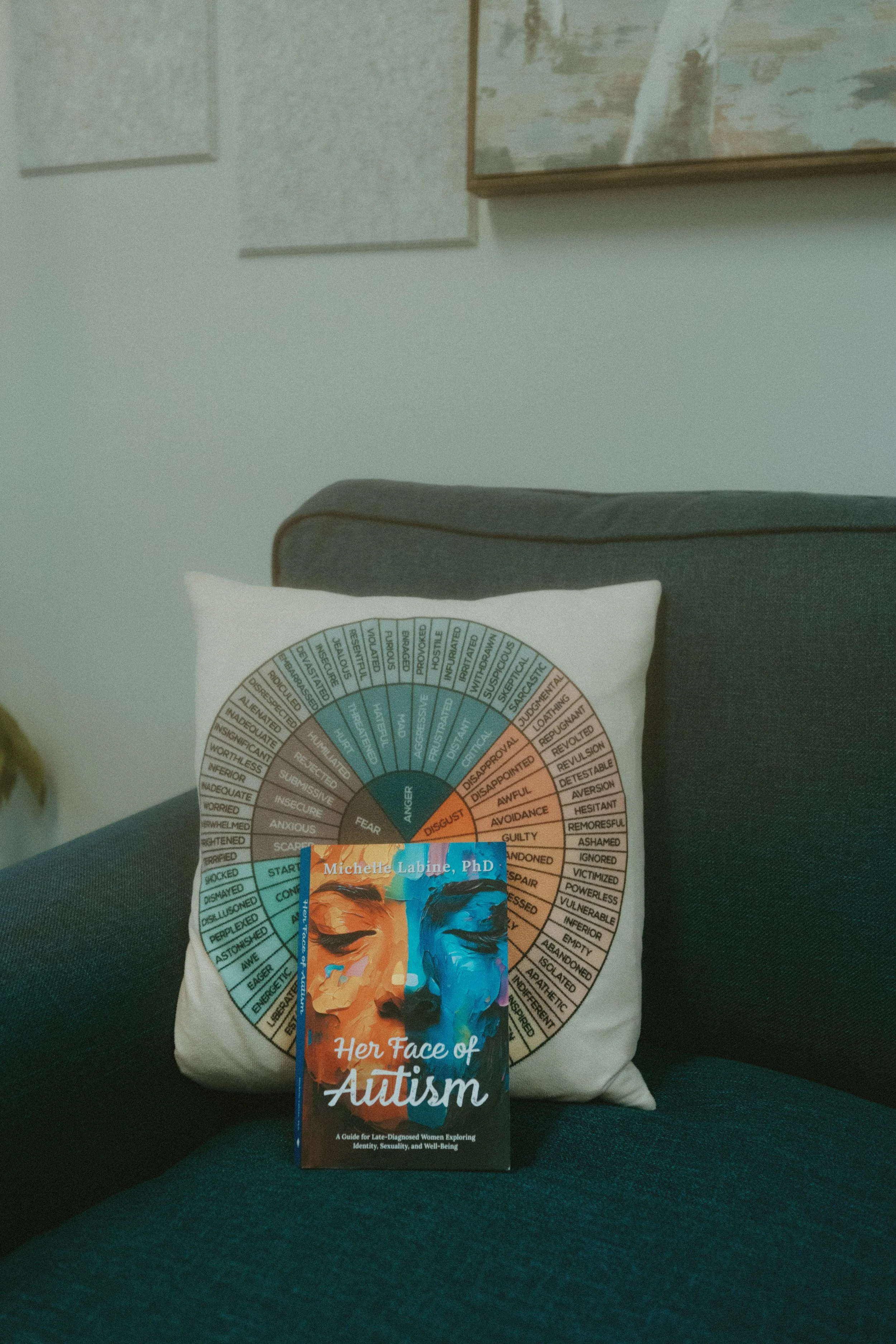 A book titled 'Her Face of Autism' by Michelle Labine, PhD, resting on a dark grey couch with a white cushion behind it. The cushion features a circular chart with various emotion labels, and a framed artwork is visible on the wall.
