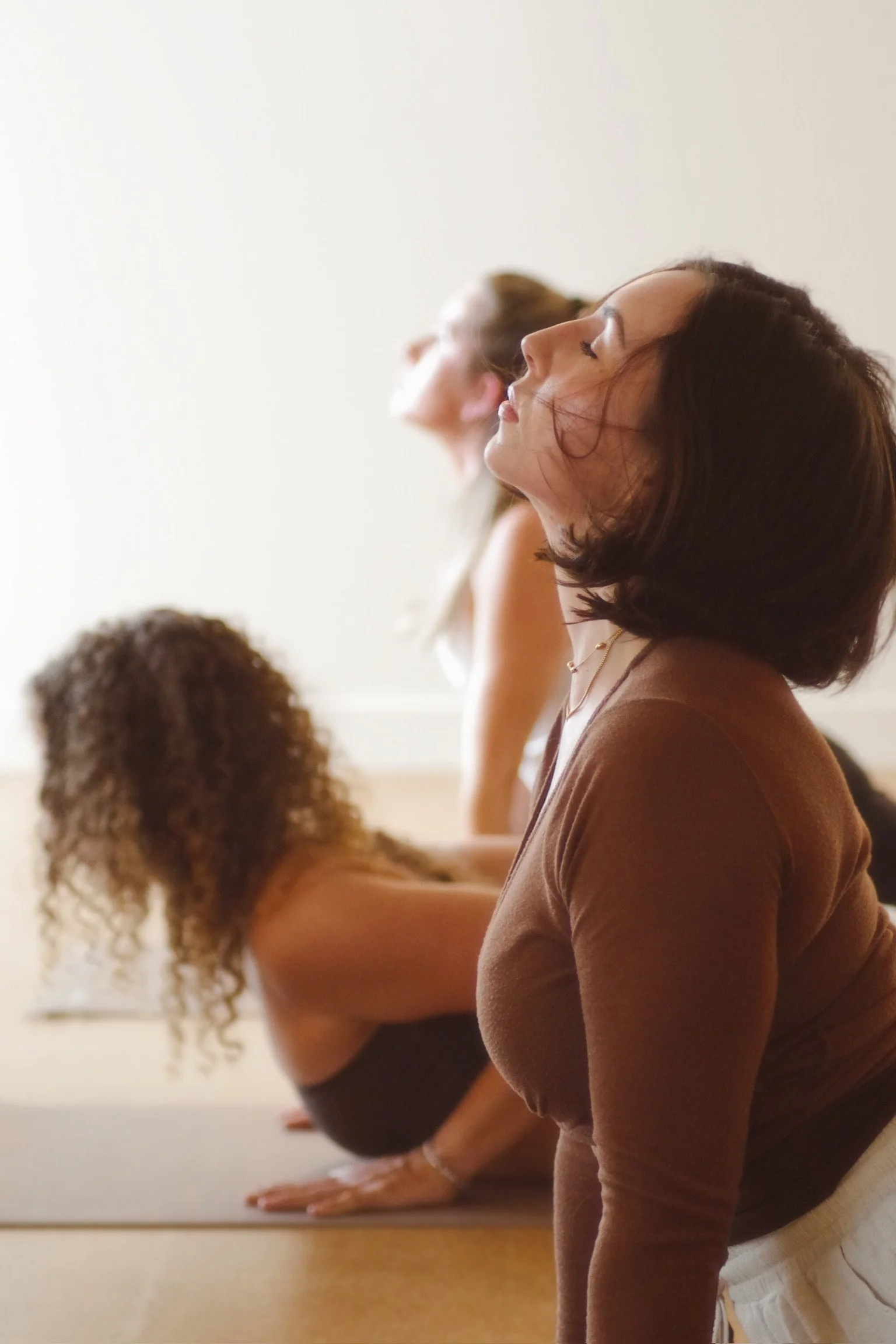 Three women practicing yoga in a studio, in a side view pose with their eyes closed and head tilted back.