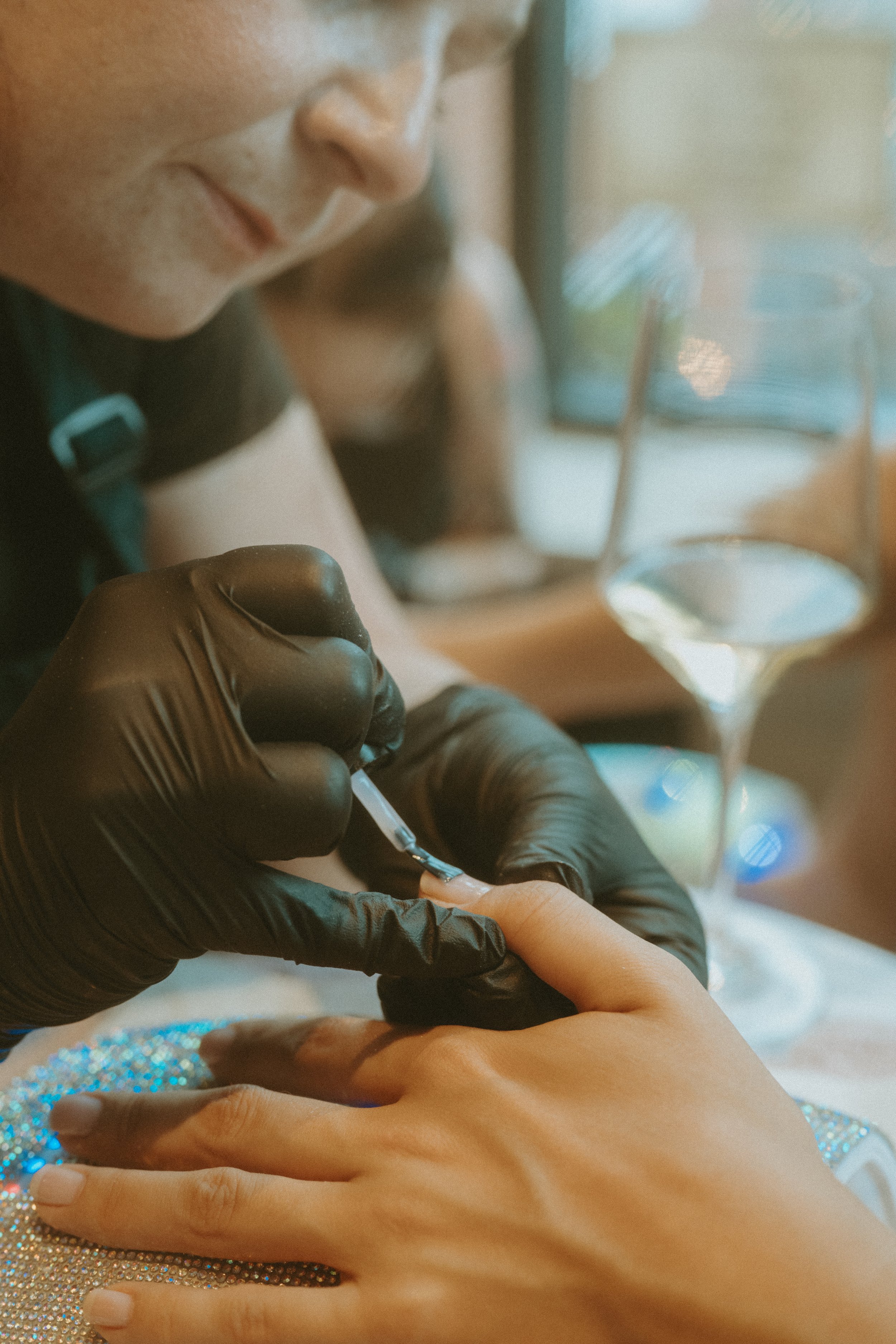 Close-up of a person receiving a manicure, with a nail technician applying nail polish. The person has their hand resting on a sparkly surface, and a glass of white wine is visible on the table.