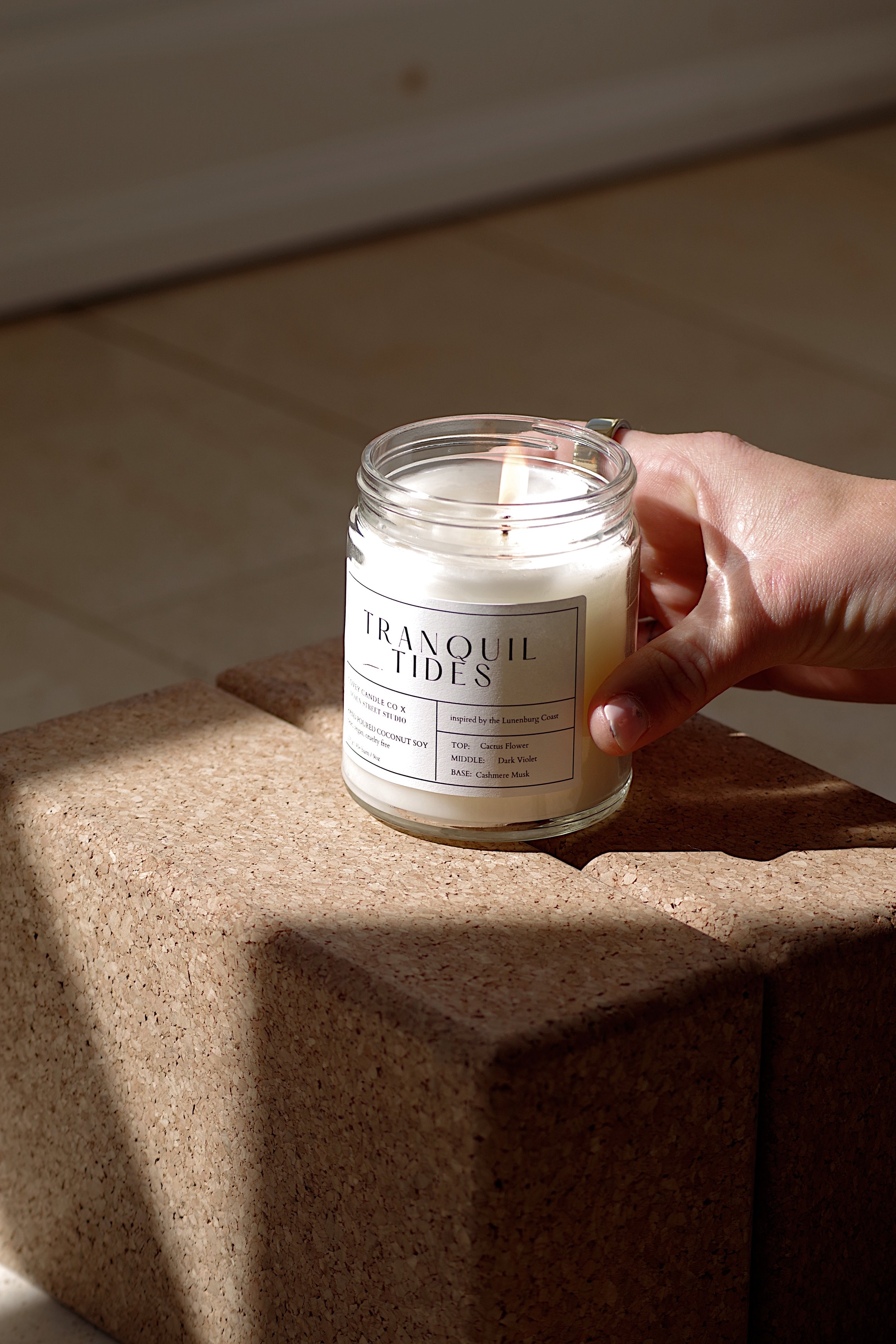 A white candle in a glass jar labeled 'Tranquil Tides,' sits on a yoga block, female hand holding candle in sunlight, yoga studio background