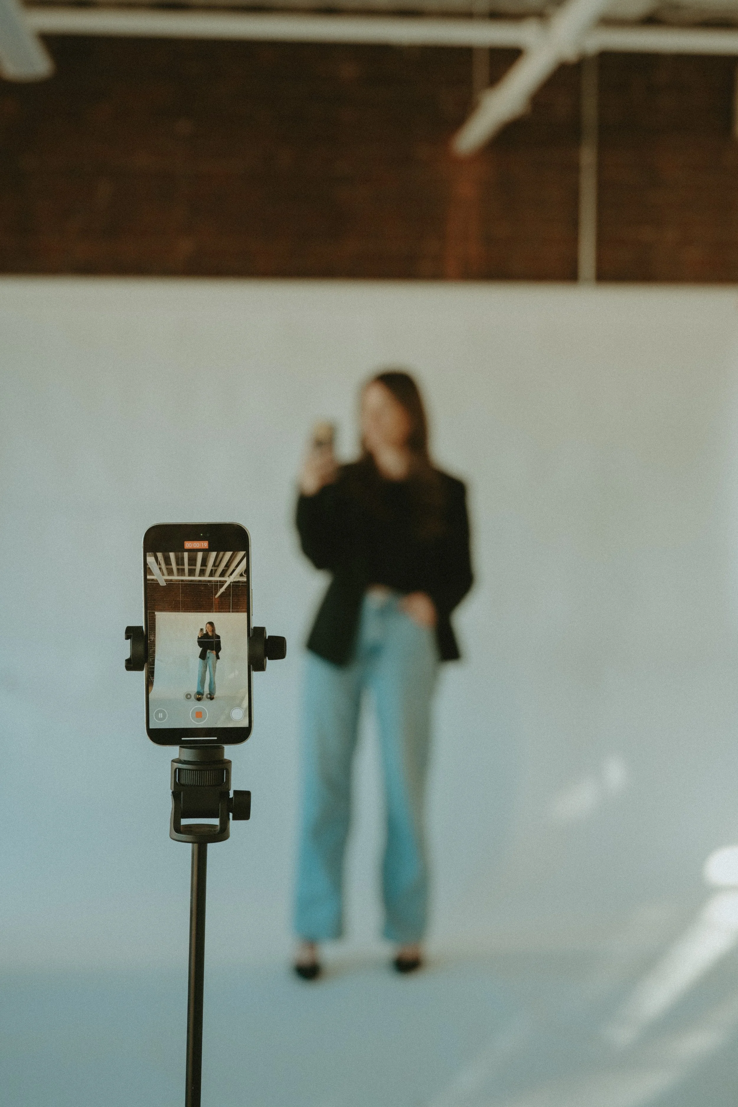 A woman in a black blazer and light blue jeans is taking a photo of herself with a smartphone mounted on a tripod in front of a plain white background, with her face blurred.