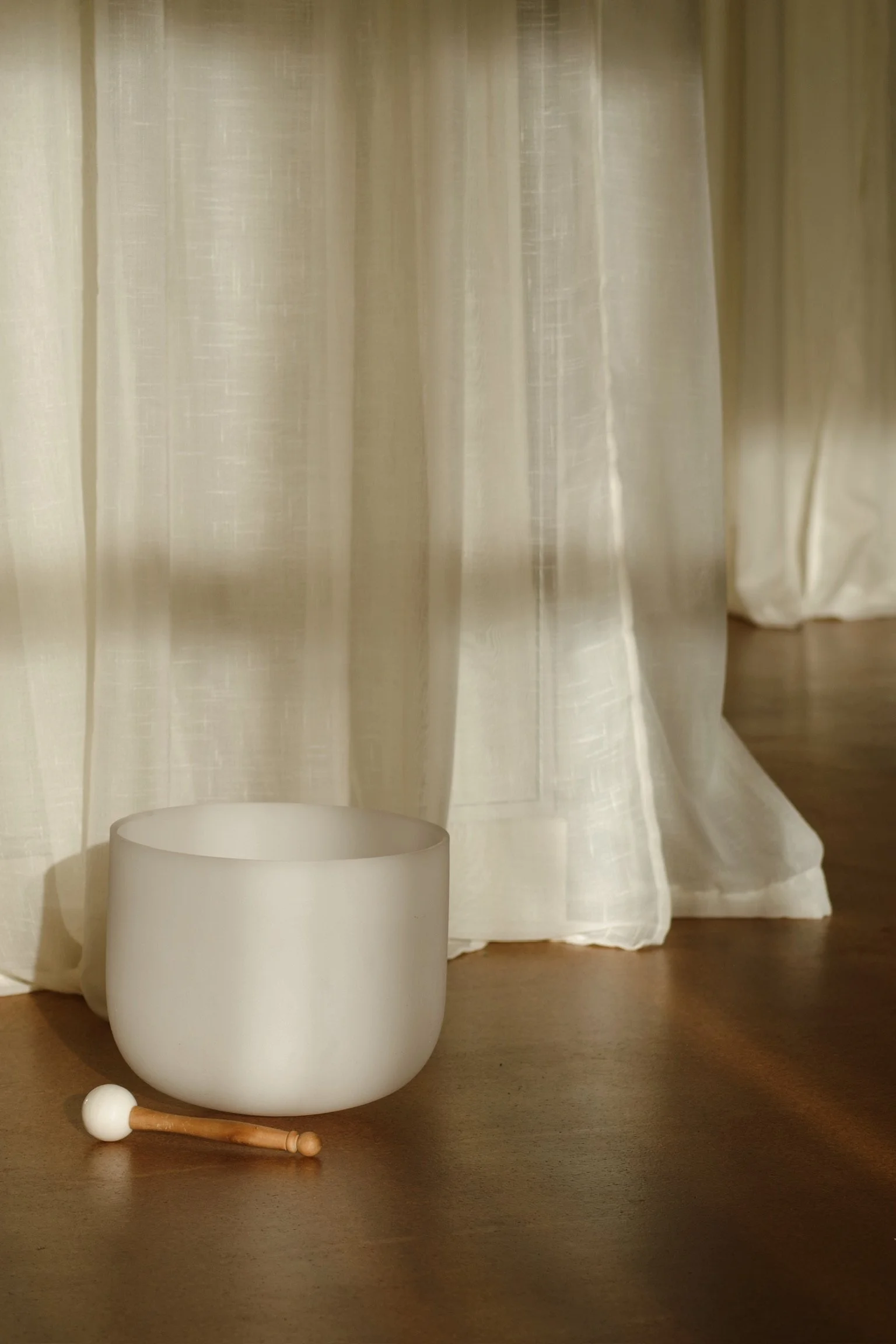 A white singing bowl with a mallet on a wooden floor next to cream-colored curtains.