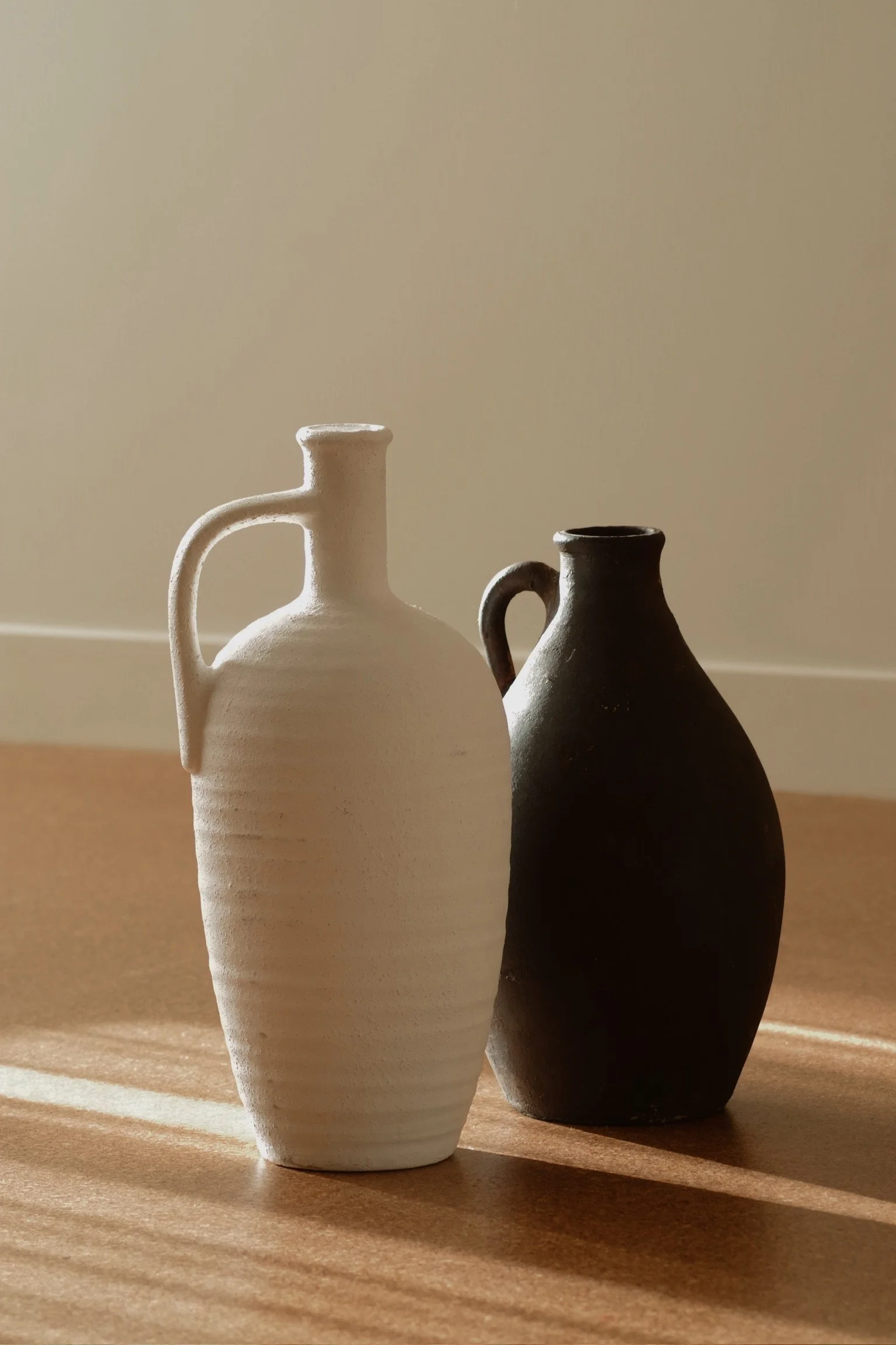 Two ceramic vases, one white and one black, positioned side by side on a wooden surface with sunlight casting shadows.