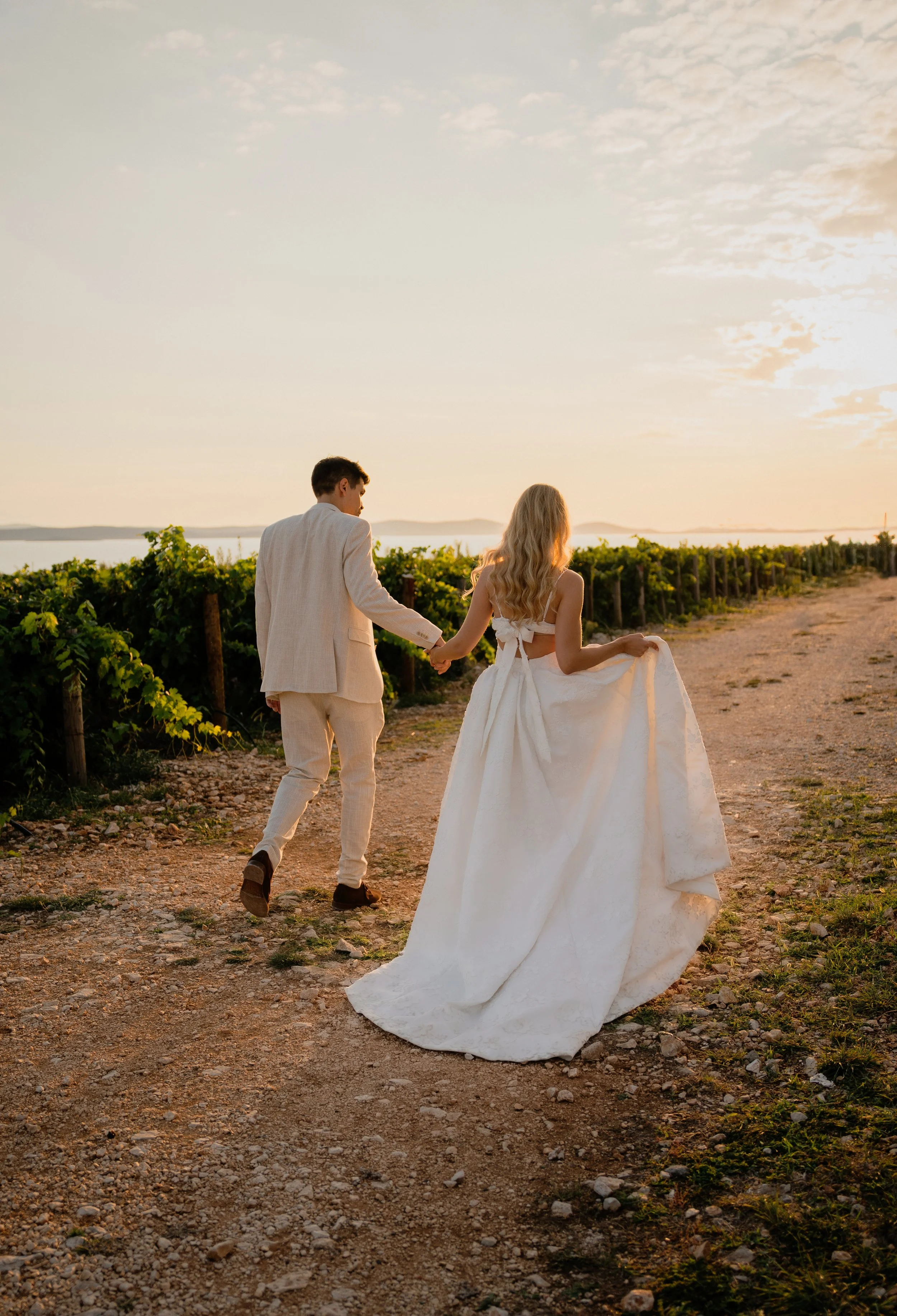 A couple walking hand in hand in a vineyard at sunset, with the woman in a white wedding dress and the man in a light-colored suit.