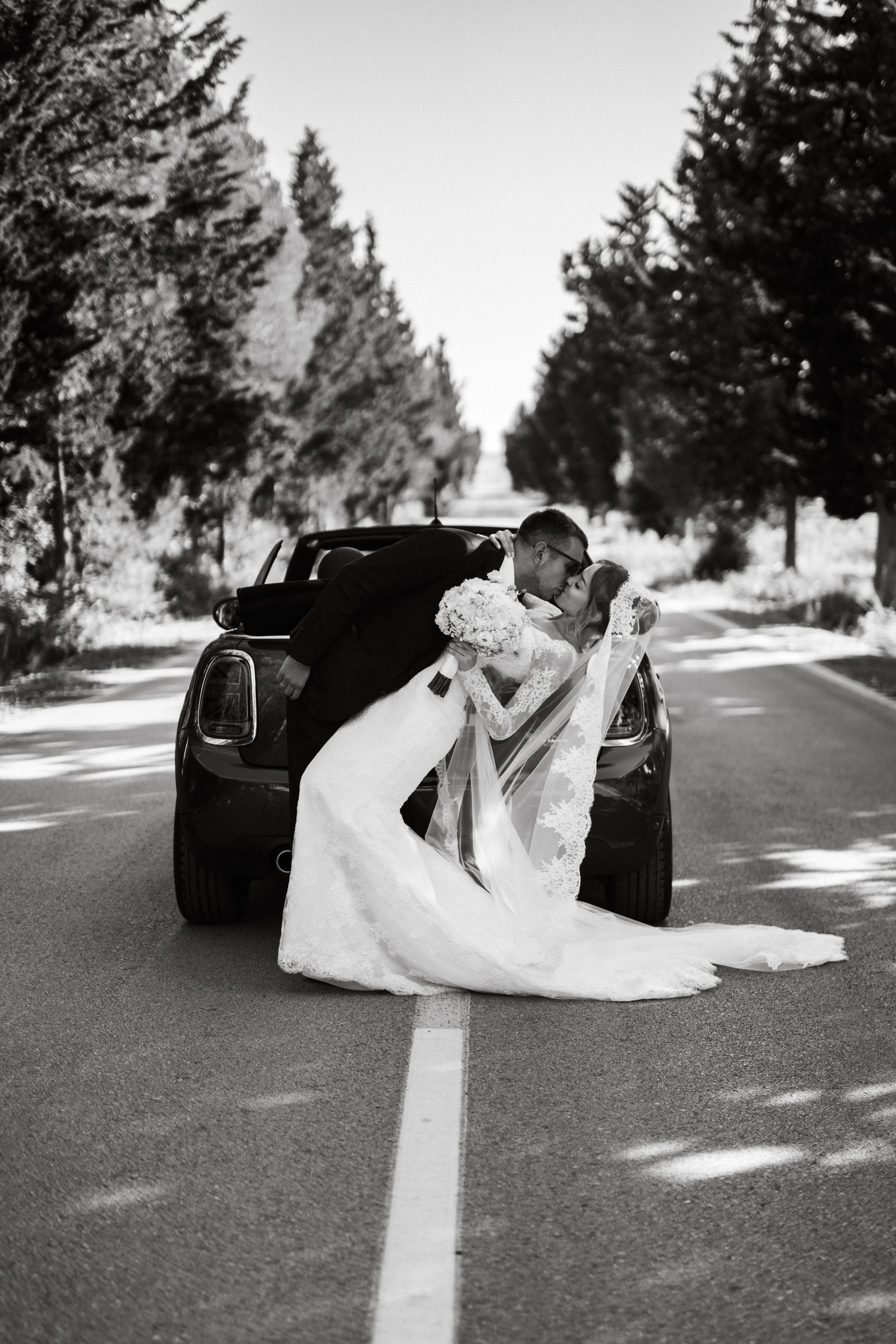 Black and white wedding photo of a bride and groom kissing, with the groom leaning over the bride who is holding a bouquet, standing in the middle of a road with trees lining both sides.